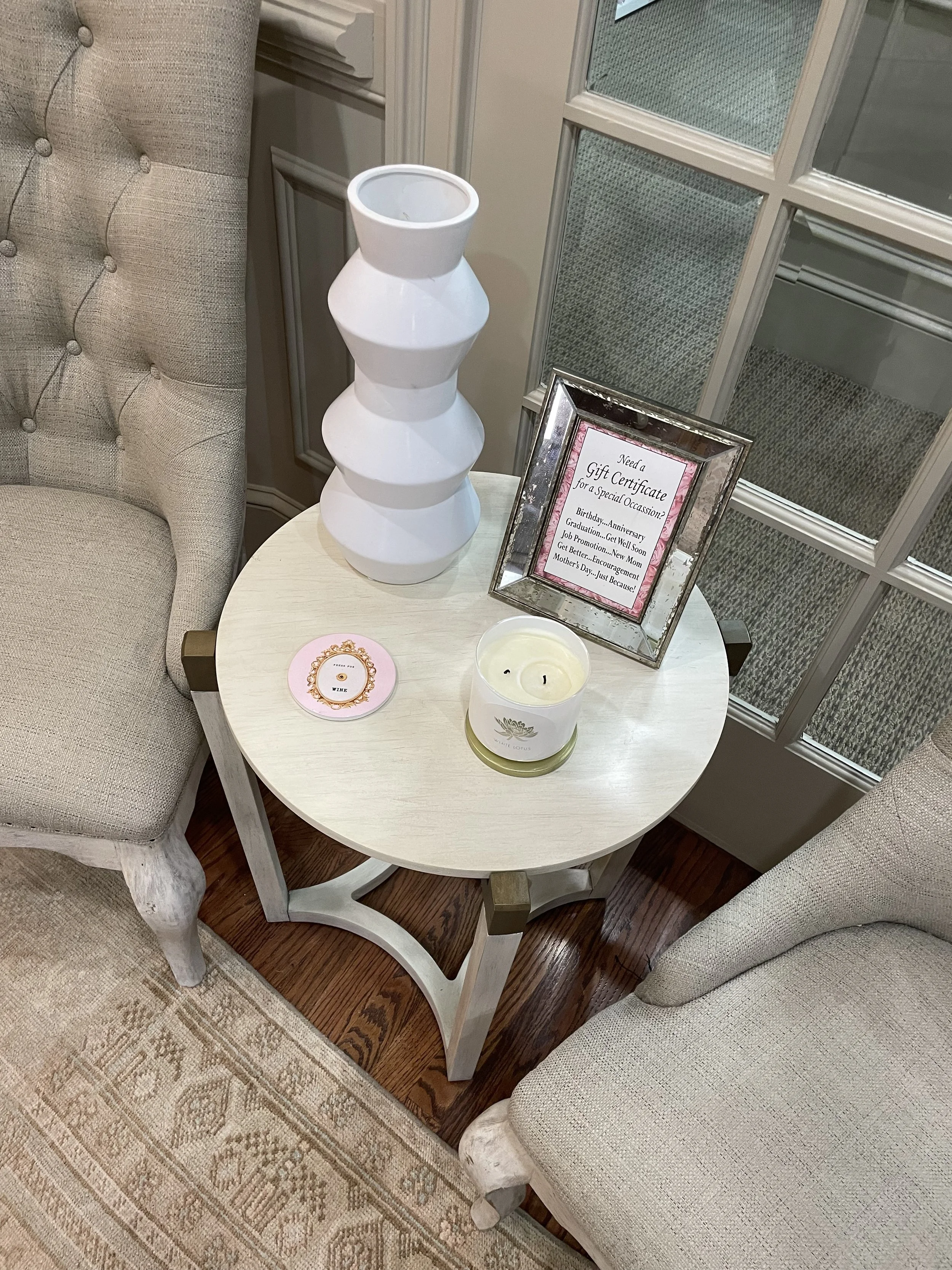 A small white round table with a large white vase, a framed sign, a lit candle, and a pink coaster, flanked by two beige upholstered armchairs, next to a window with glass panes.