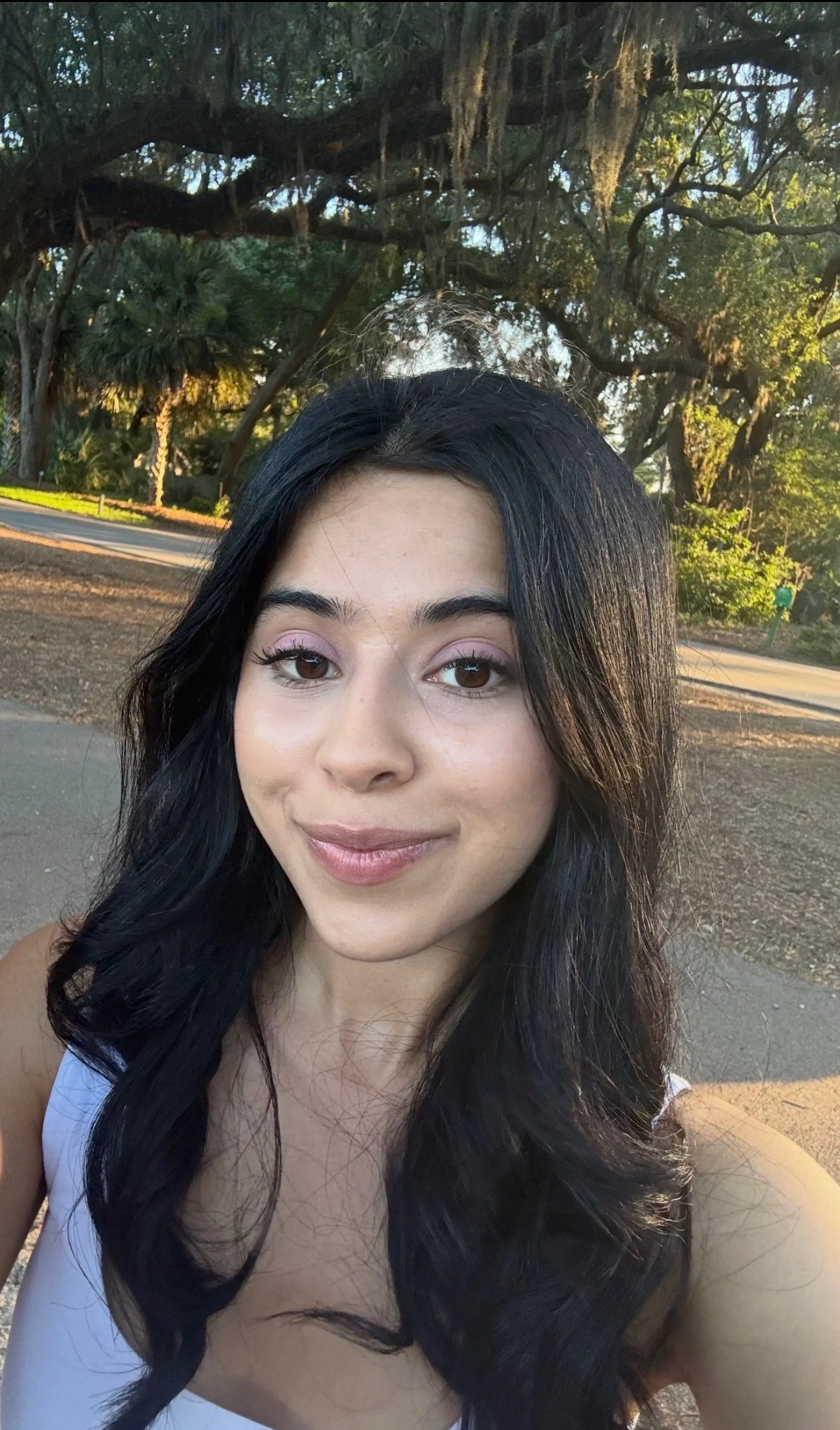 Close-up of a young woman with long dark hair and light makeup, smiling outdoors in a park with trees and a pathway in the background.