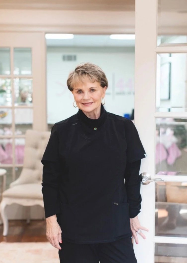 A smiling woman with short light brown hair, wearing a black top and earrings, standing in a cozy, well-lit room with white walls and furniture.