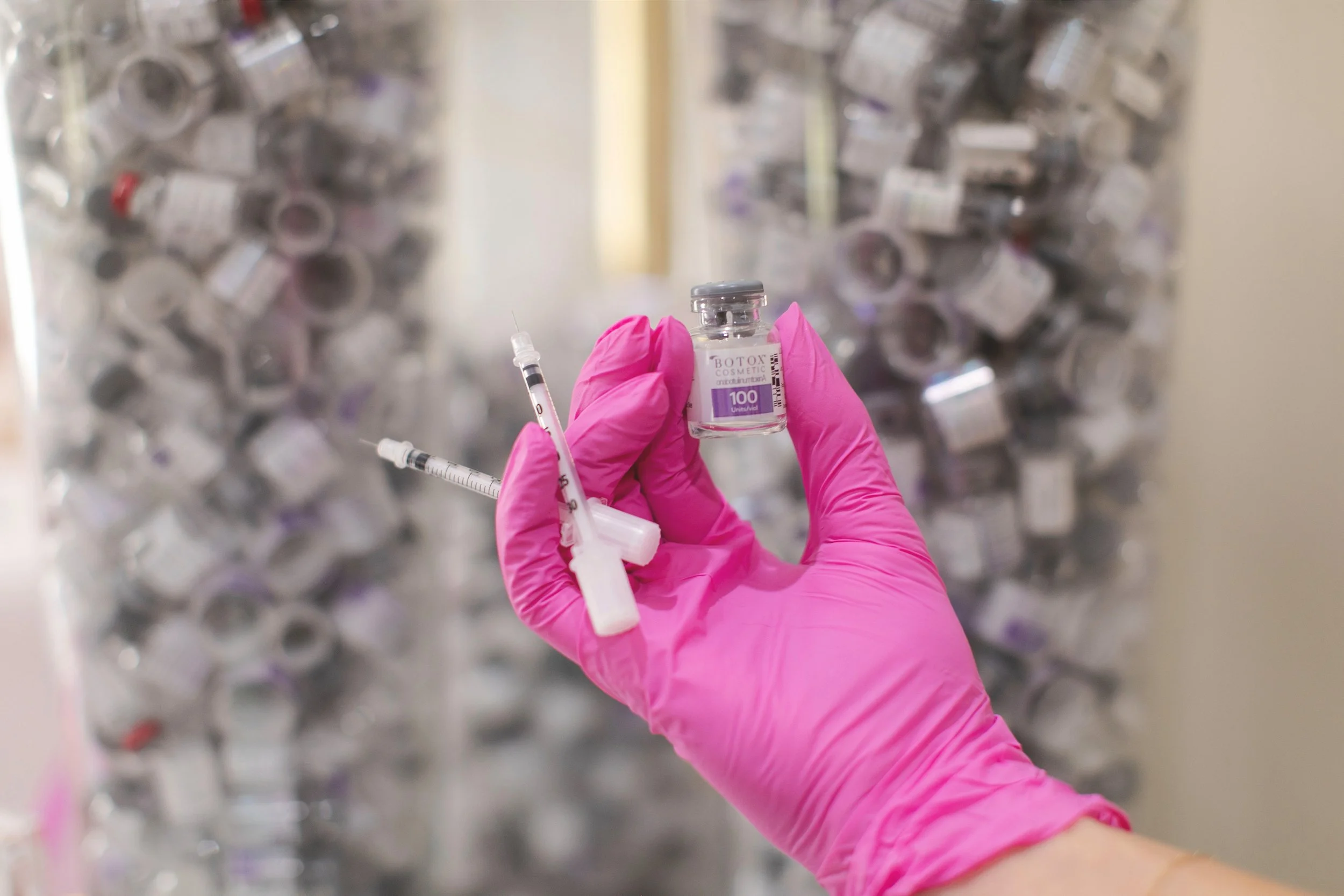 A hand wearing a pink glove holds a small vial labeled 'Botox' and two syringes against a background of numerous similar vials and syringes.