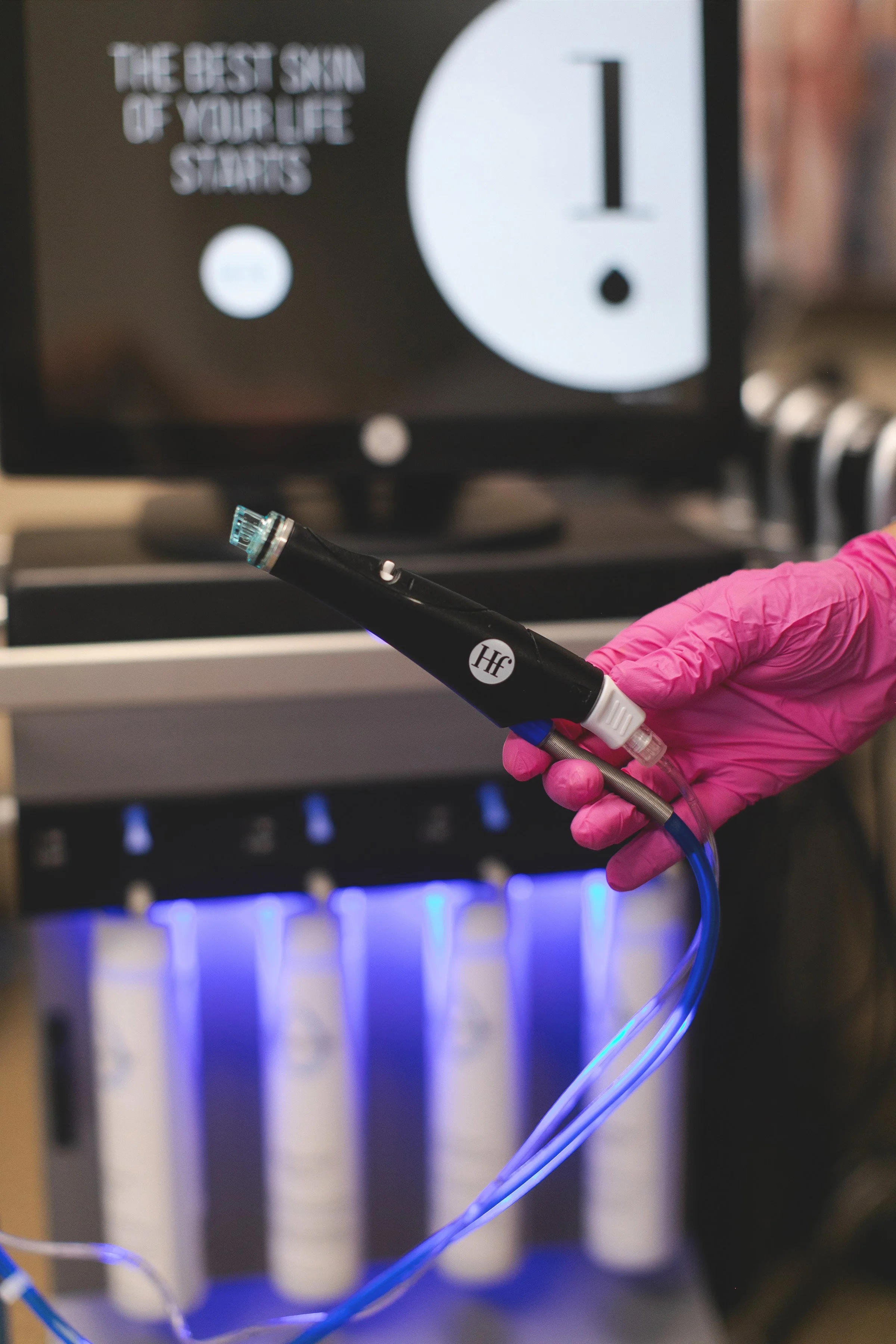 A person wearing pink gloves holding a black dermatology or medical device connected to blue tubes with a medical machine in the background.