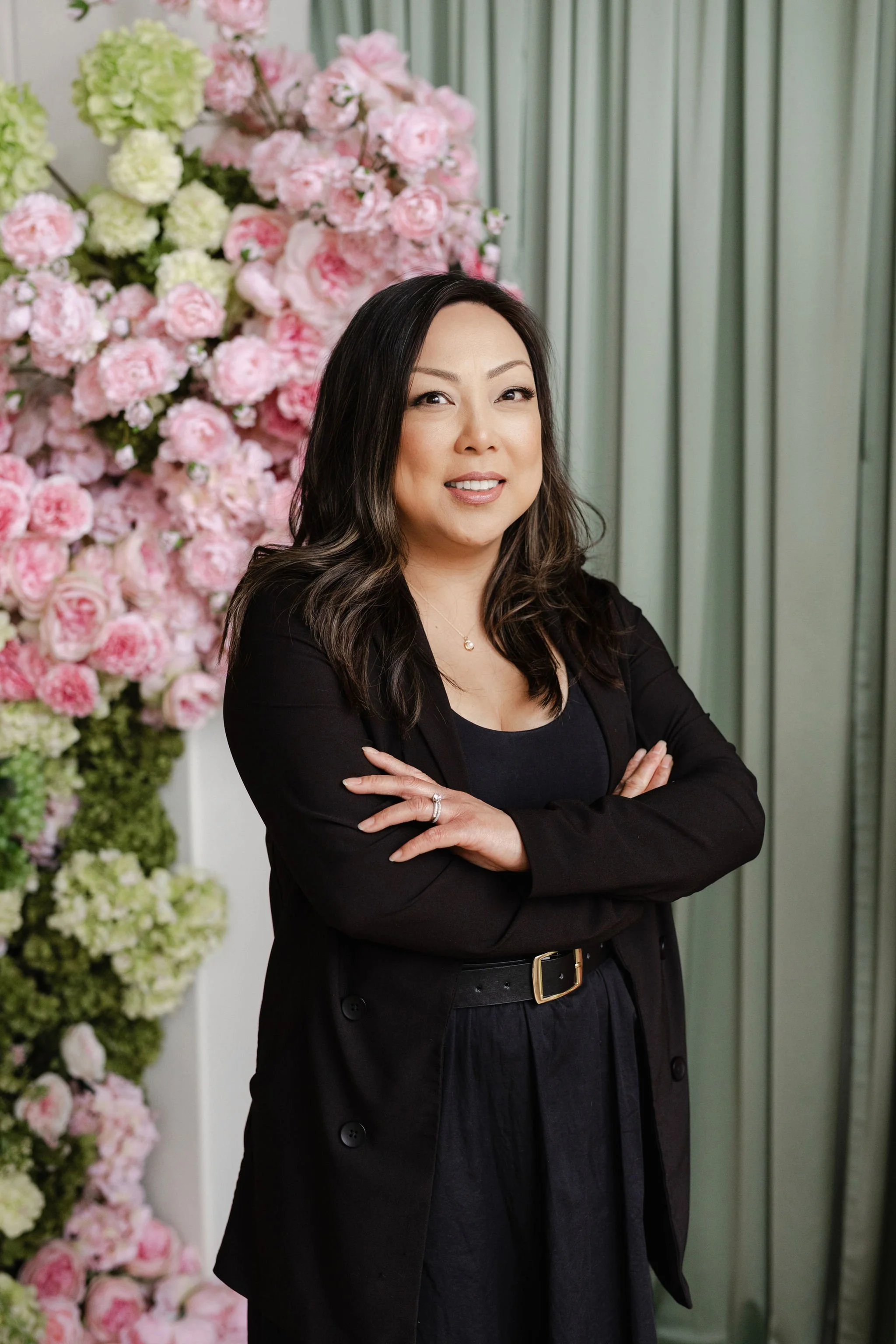 Jennifer, in a black dress, is arranging a dining table with tall glass candle holders, pink wine glasses, and a floral centerpiece.