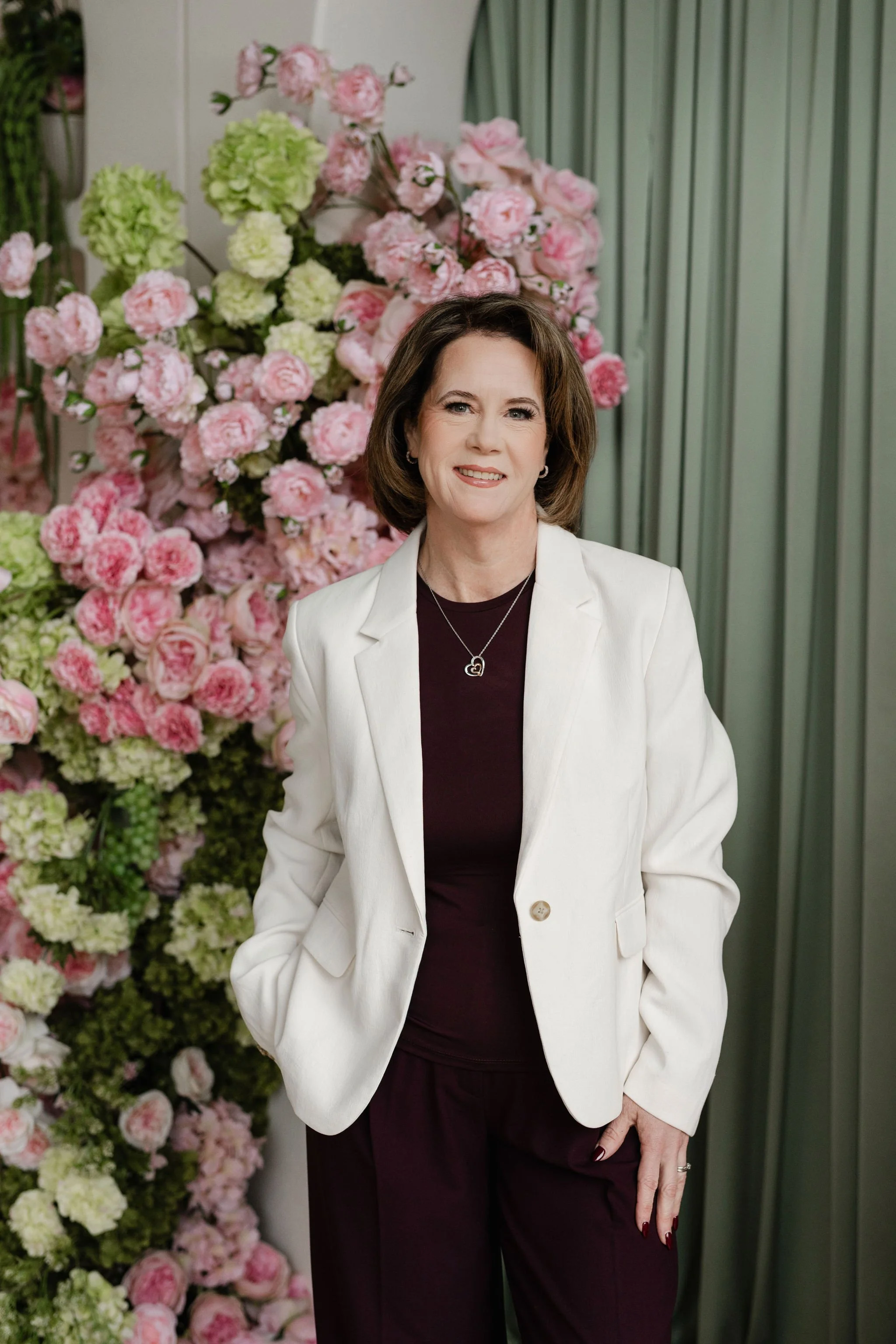 Vicky is in a pink blazer and floral top, standing in front of decorated shelves.