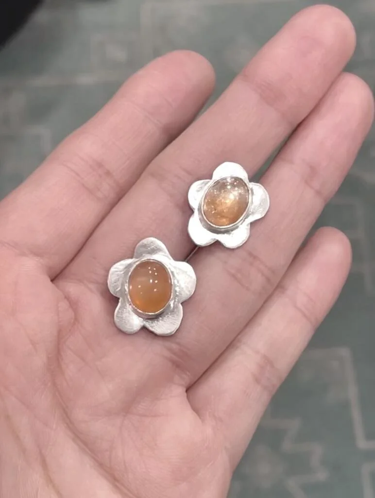 A hand holding a pair of silver earrings with flower-shaped designs and oval orange sunstone and moonstone in the center of each