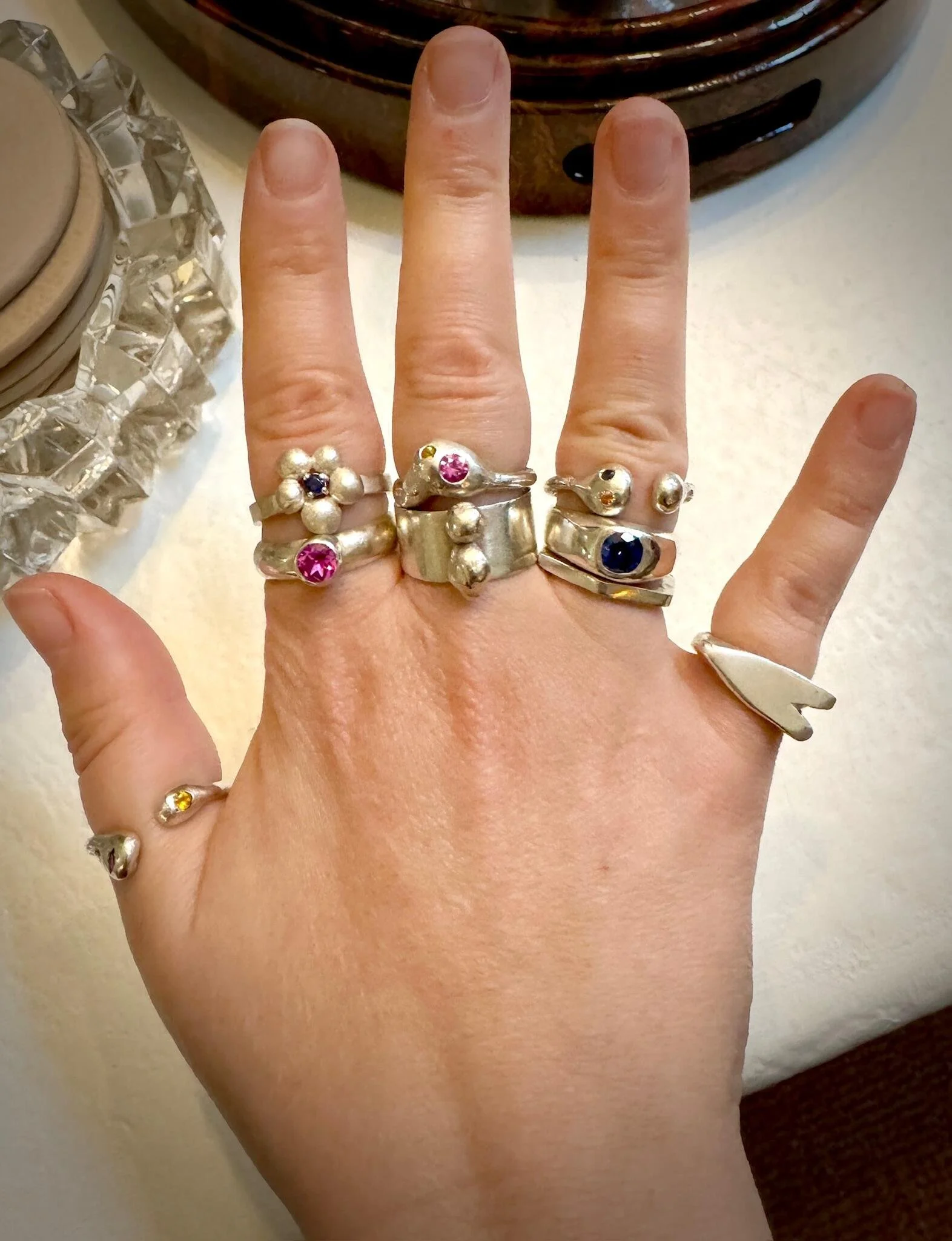 A person's hand wearing nine silver rings, including a flower-shaped ring with a pink gemstone, a ring with a large blue gemstone, a simple band with two silver balls, and a elongated heart-shaped ring