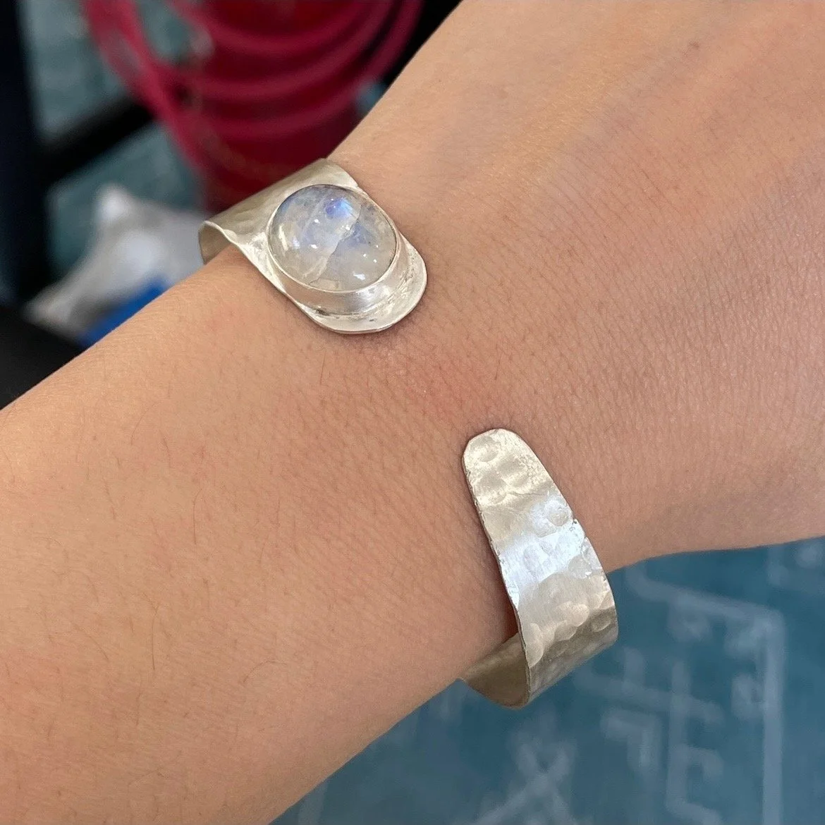 A person's wrist adorned with a silver bracelet with a large, round moonstone set on one end, against a blurred background.