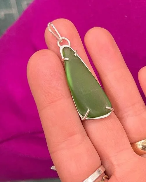 A hand holds a pendant with a piece of green sea glass set in a pronged silver frame, against a fuscia background