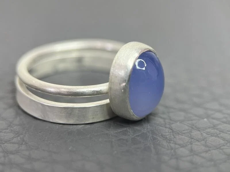A silver ring with a blue oval gemstone set in a bezel setting sitting on top of a simple silver band