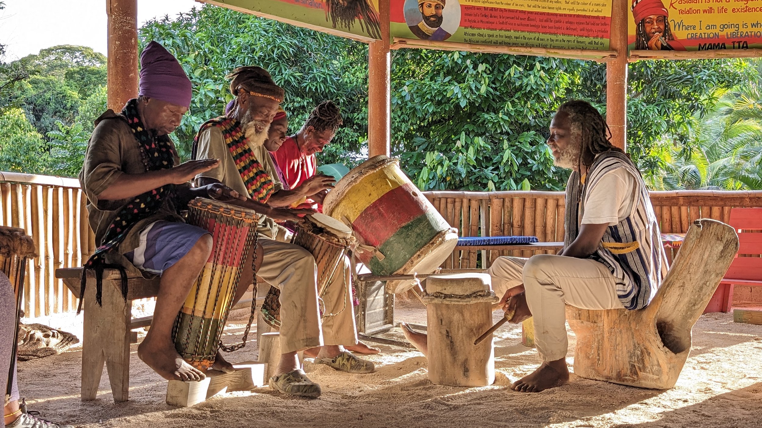 A group of five people sitting under a covered outdoor area with lush green trees in the background. They are engaged in playing traditional drums and percussion instruments while sitting on wooden stools or logs, appearing to be part of a musical or cultural gathering.