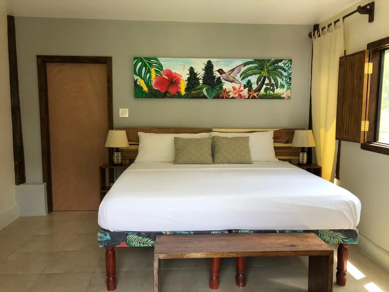 Hotel room with a bed, two pillows, two bedside lamps, wooden furniture, a tropical artwork above the bed, and open windows with yellow curtains.
