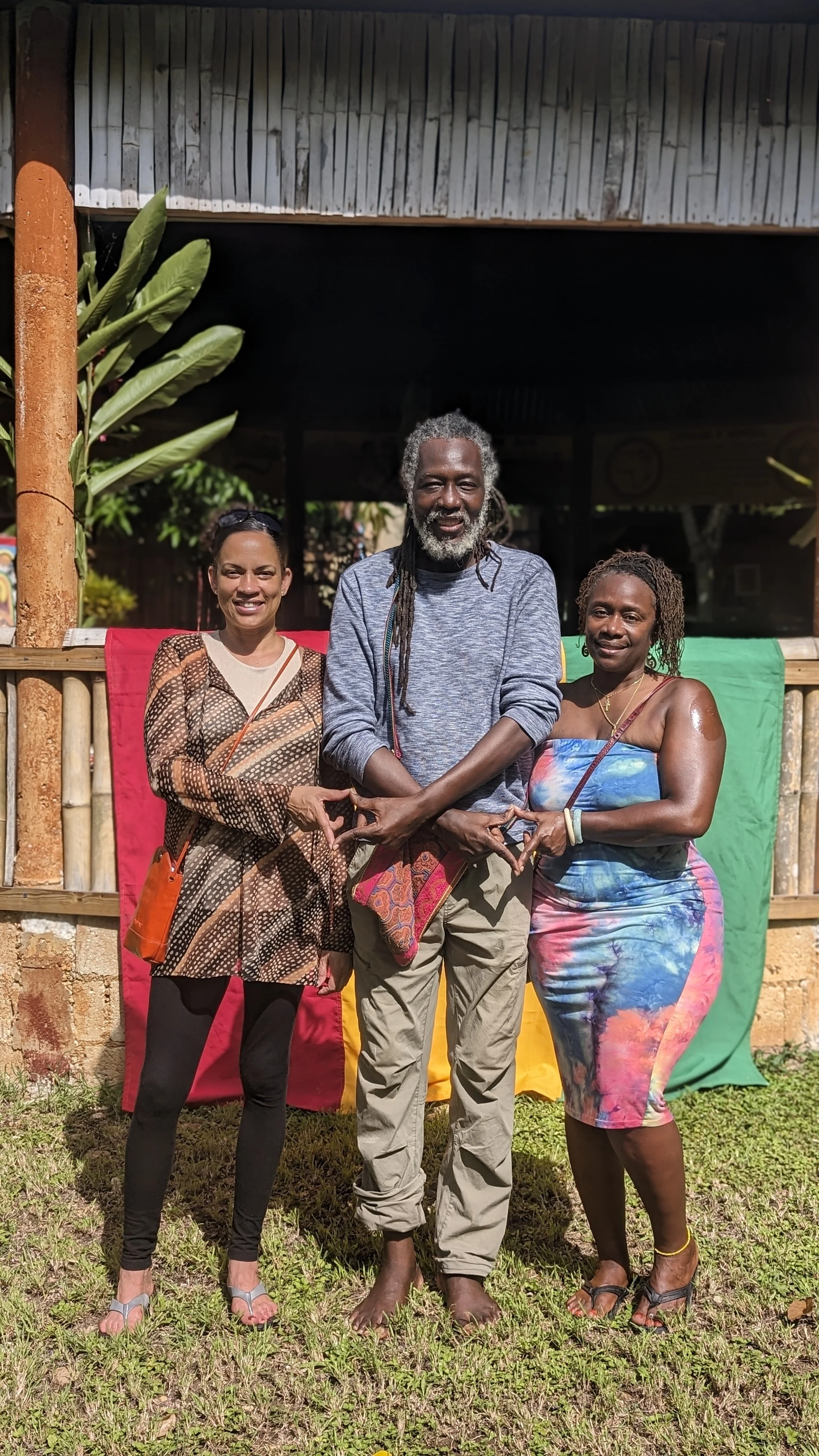 Three people standing together outdoors in front of a colorful backdrop, smiling and forming a heart shape with their hands.