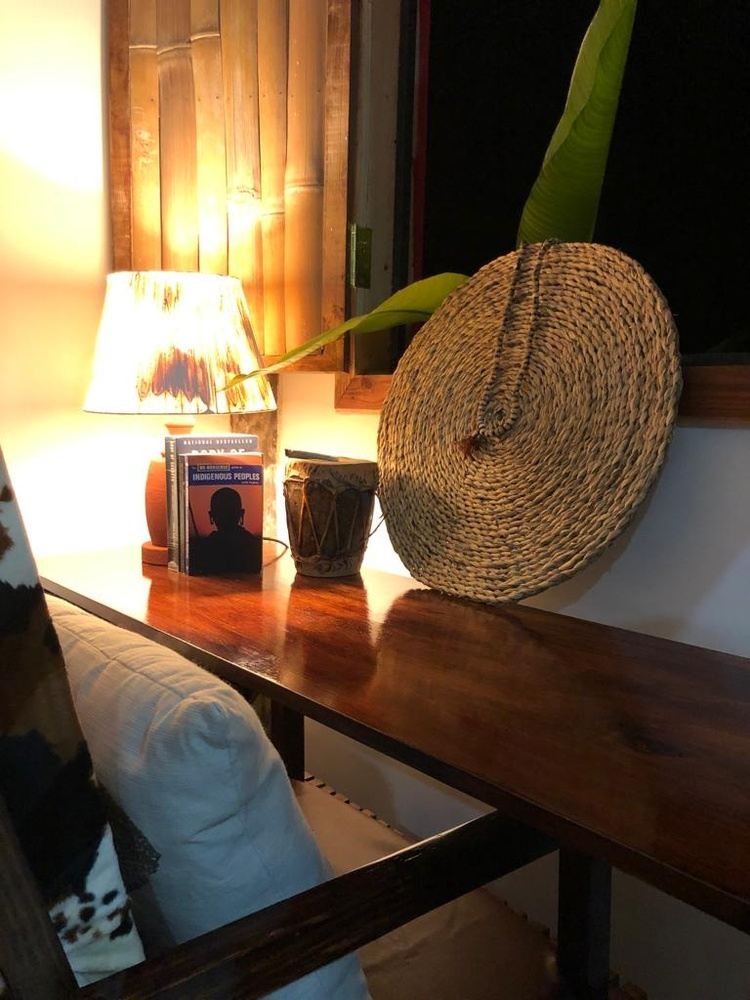 Indoor wooden desk with a lamp, a book titled 'Indigenous Peoples,' a small drum, and a woven circular wall decoration. Part of a white pillow and a plant leaf are also visible.