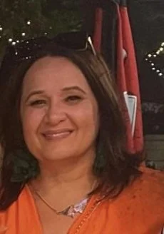 A woman smiling with shoulder-length brown hair, wearing an orange top and makeup, with a background of festoon lights and red umbrellas.
