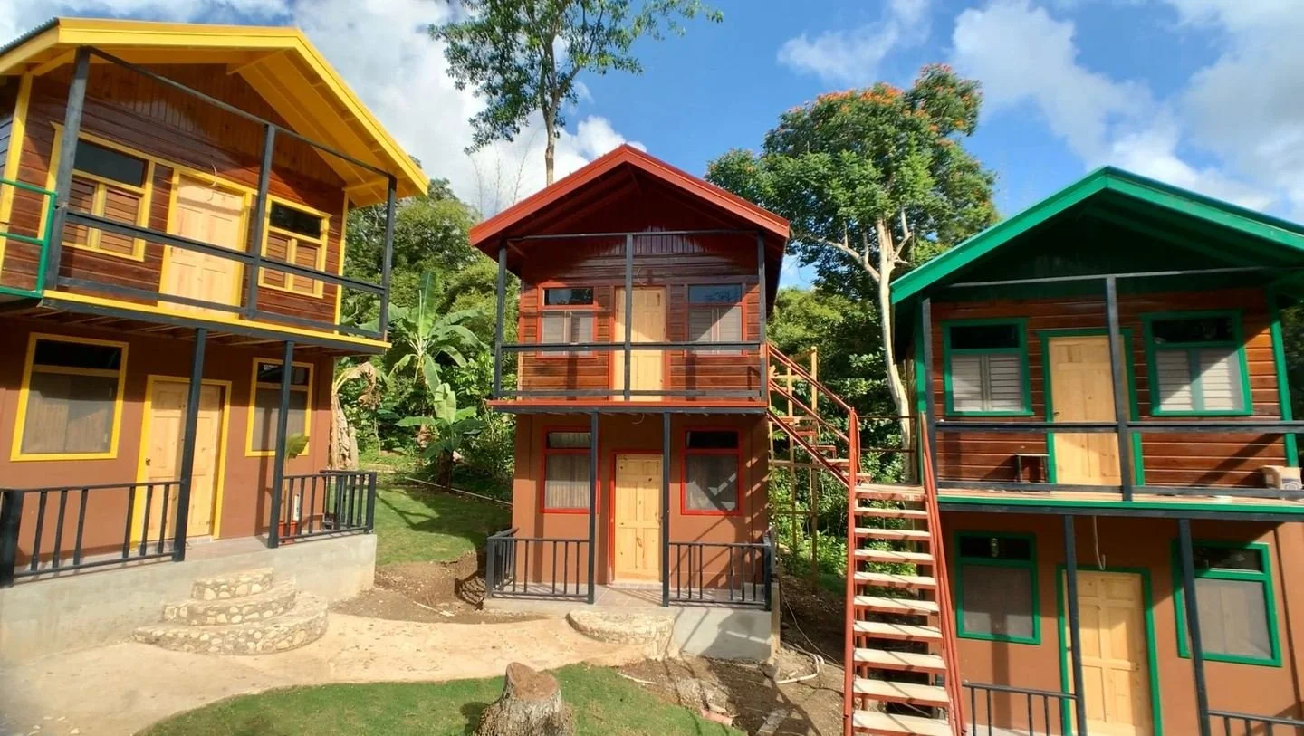 Small colorful houses with wood exteriors, each with a door and windows, on a grassy area with trees in the background under a partly cloudy sky.