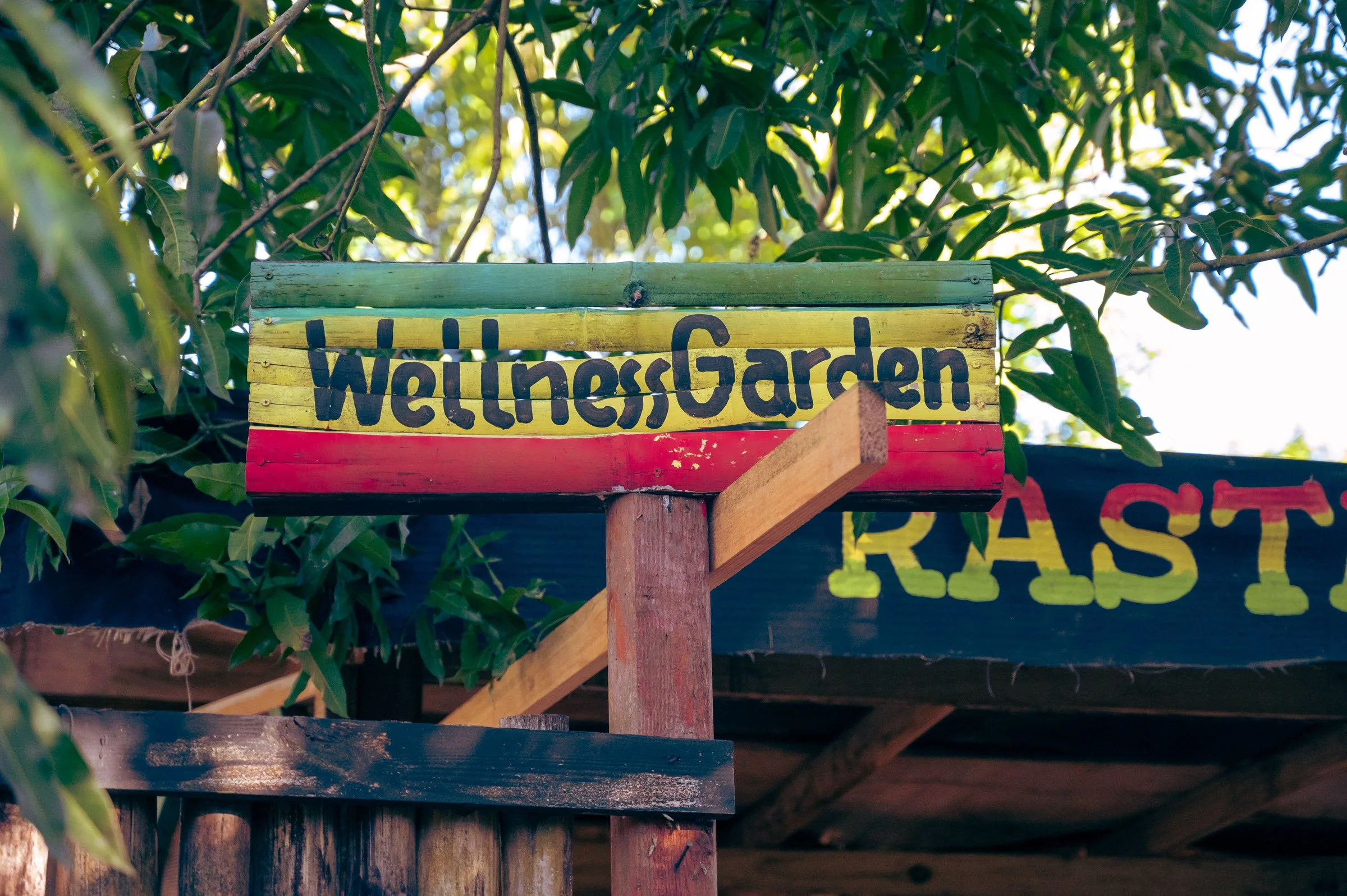 Colorful sign reading 'Wellness Garden' mounted on a wooden post, under tree with green leaves in the background.