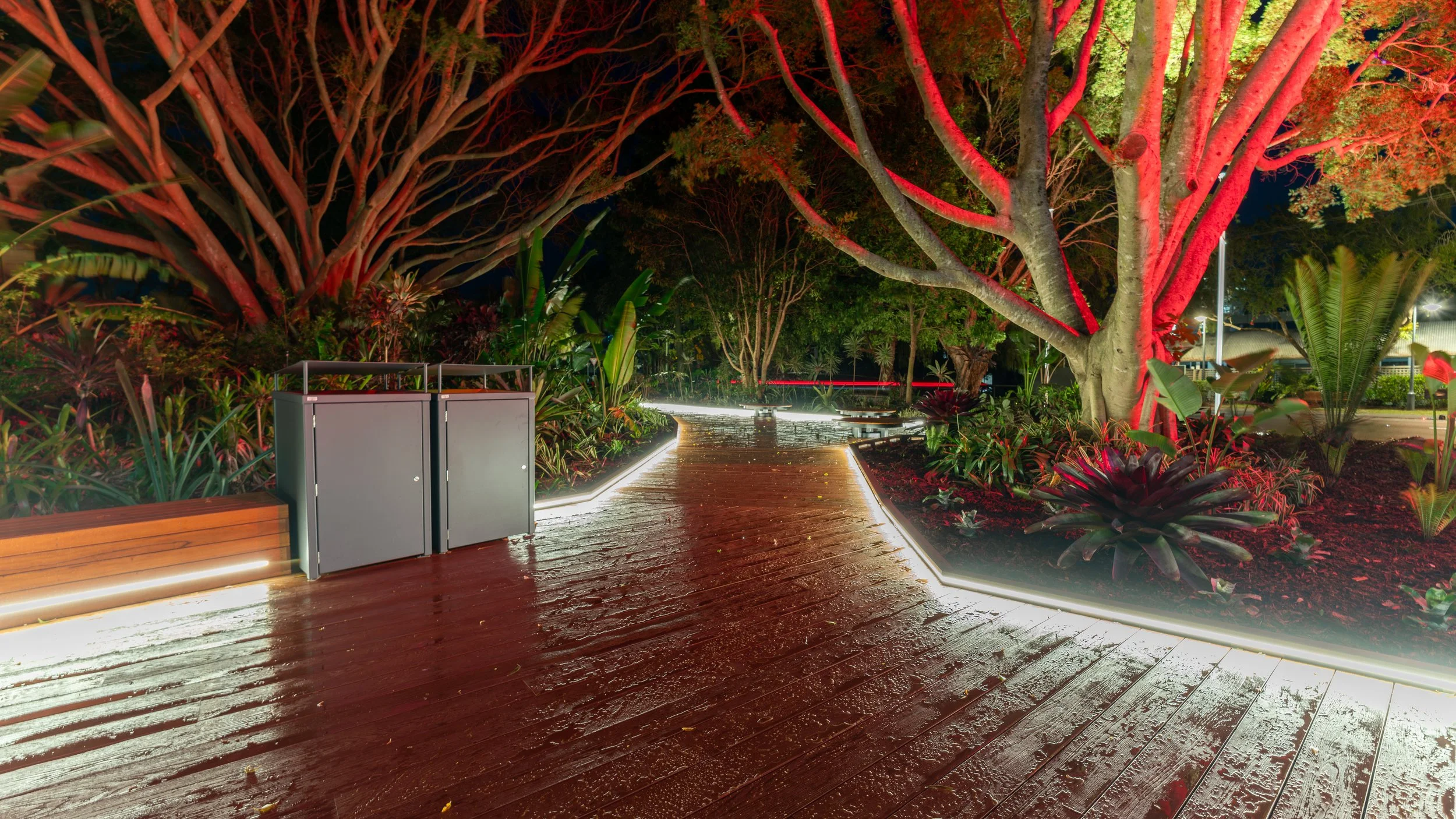 Night view of a wet wooden pathway illuminated with strip lighting, surrounded by lush green plants and large trees with branches highlighted in red lights.