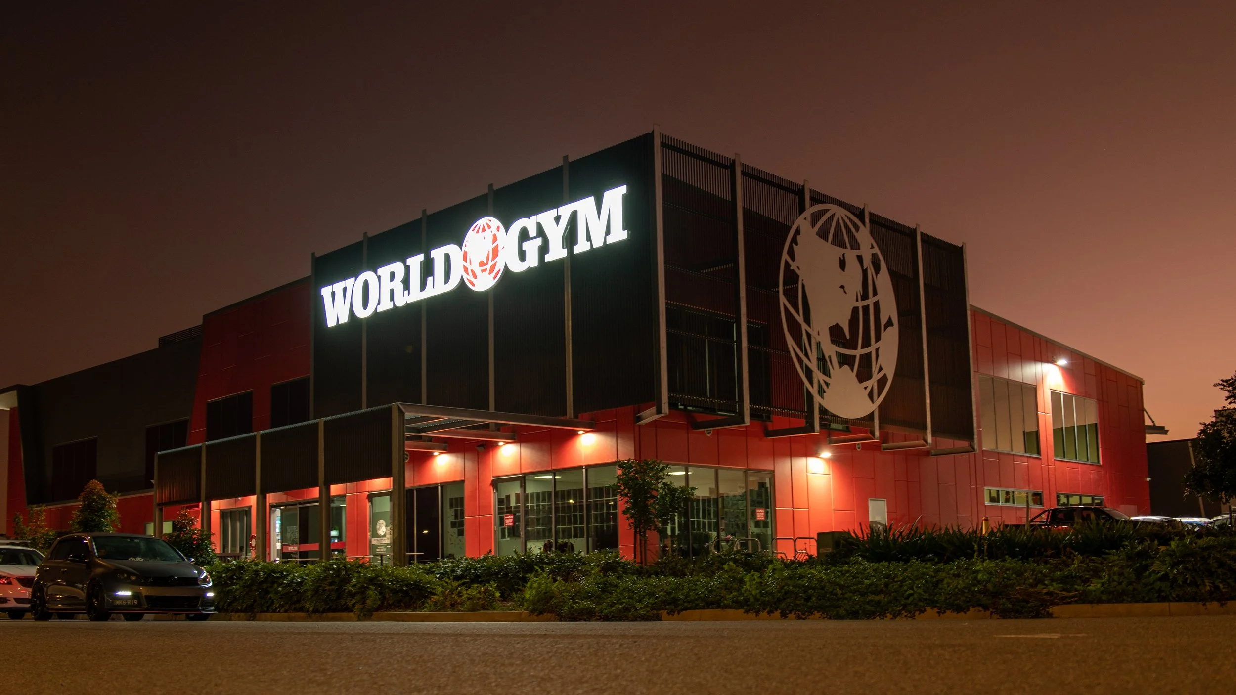 Night view of the exterior of a building with the sign “World Gym” illuminated, featuring a globe logo, with parked cars in front and a dark sky in the background.