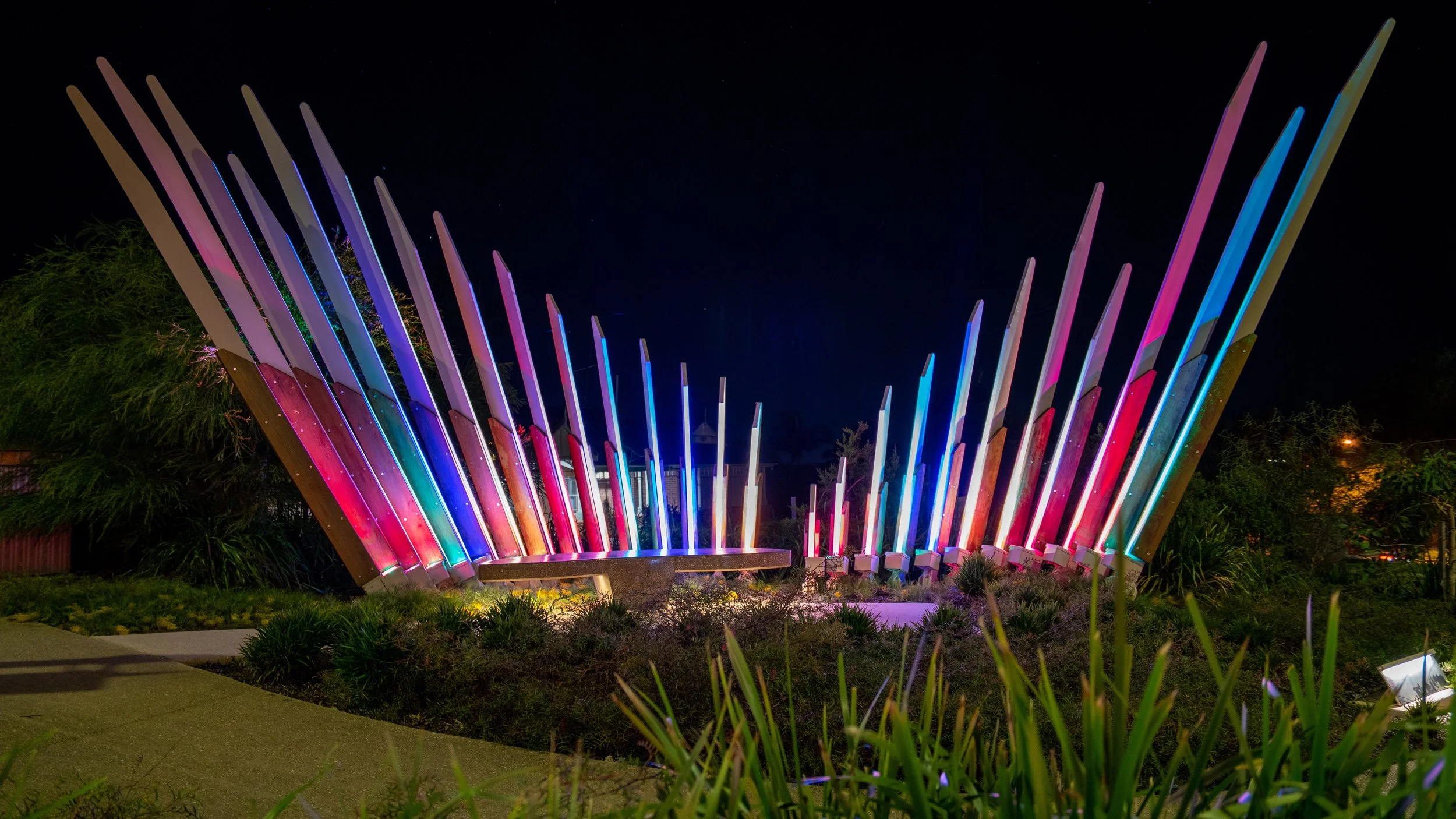 A colorful outdoor art installation at night featuring illuminated vertical poles arranged in a semi-circle, with vibrant rainbow colors shining against a dark sky, surrounded by greenery and a concrete pathway.