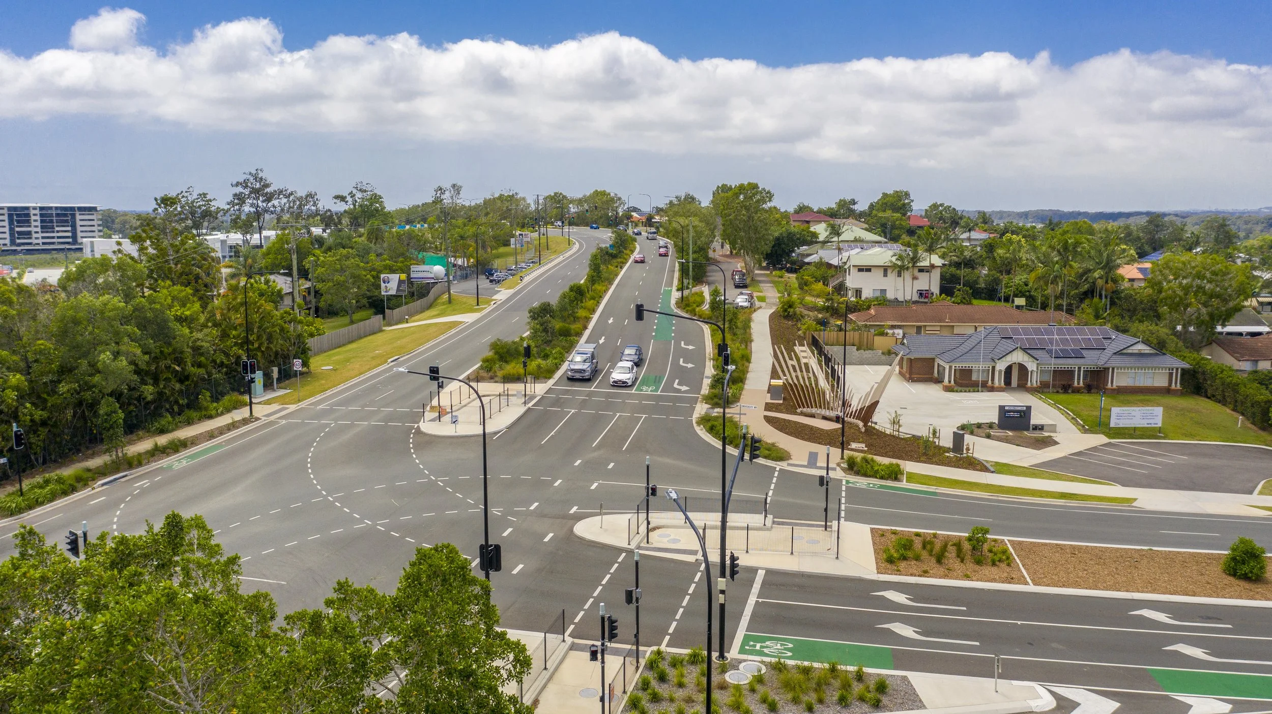 Aerial view of a busy intersection with traffic lights, crosswalks, and several cars on the road, surrounded by residential houses, trees, and a partly cloudy sky.