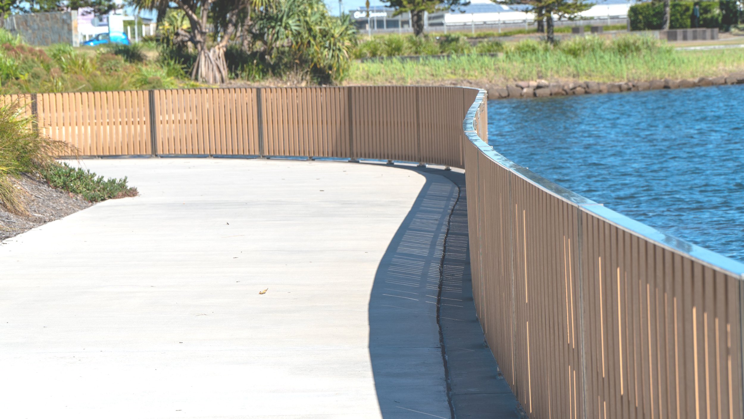 A curved concrete walkway alongside a body of water, bordered by a wooden fence on one side and greenery in the background.