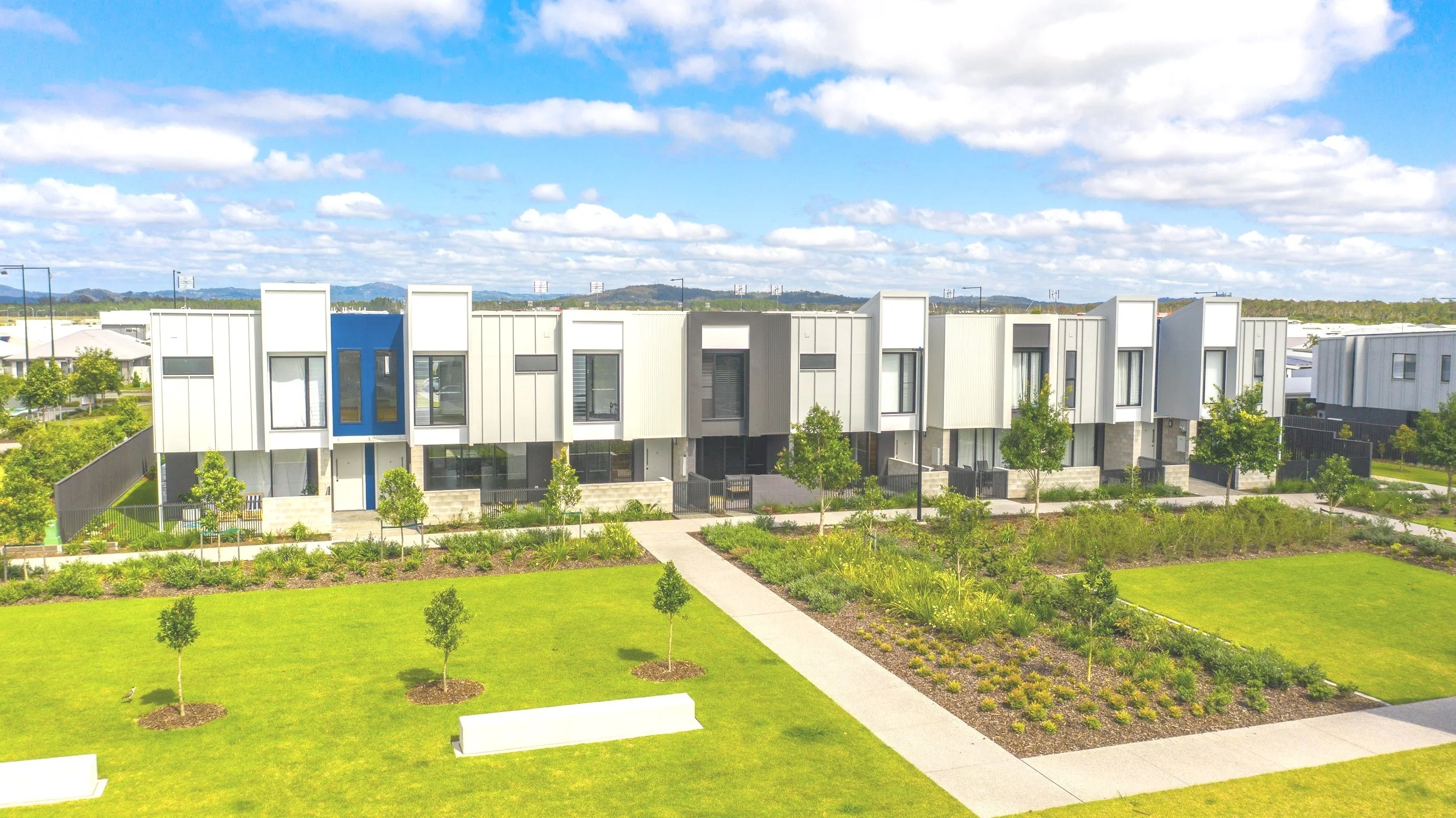 Modern row of townhouses with contemporary architecture, small trees, landscaped garden, and walking paths in a suburban neighborhood under a partly cloudy sky.
