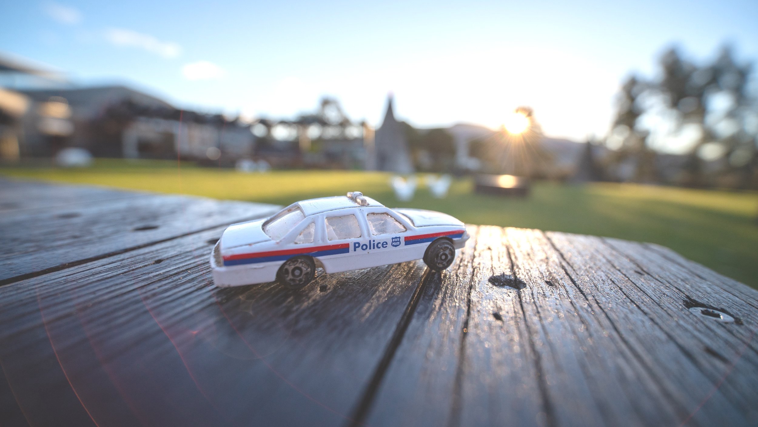 A small toy police car on a wooden surface outdoors with a grassy area and buildings in the background, sunlight shining in the distance.