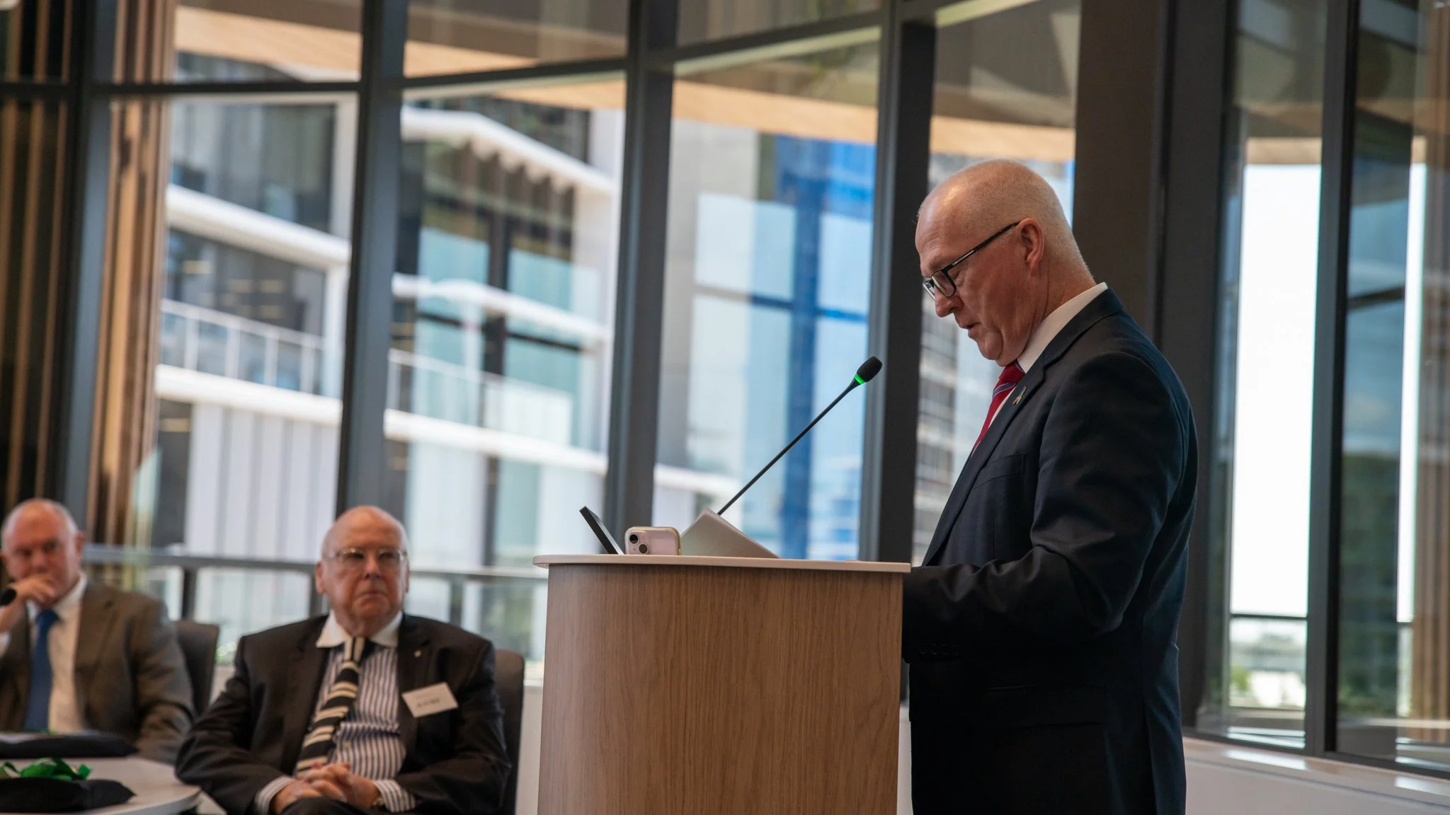 A man in a suit and tie is standing at a lectern, reading from a paper, with a microphone in front of him. In the background, seated men are watching, with large windows showing a modern cityscape outside.
