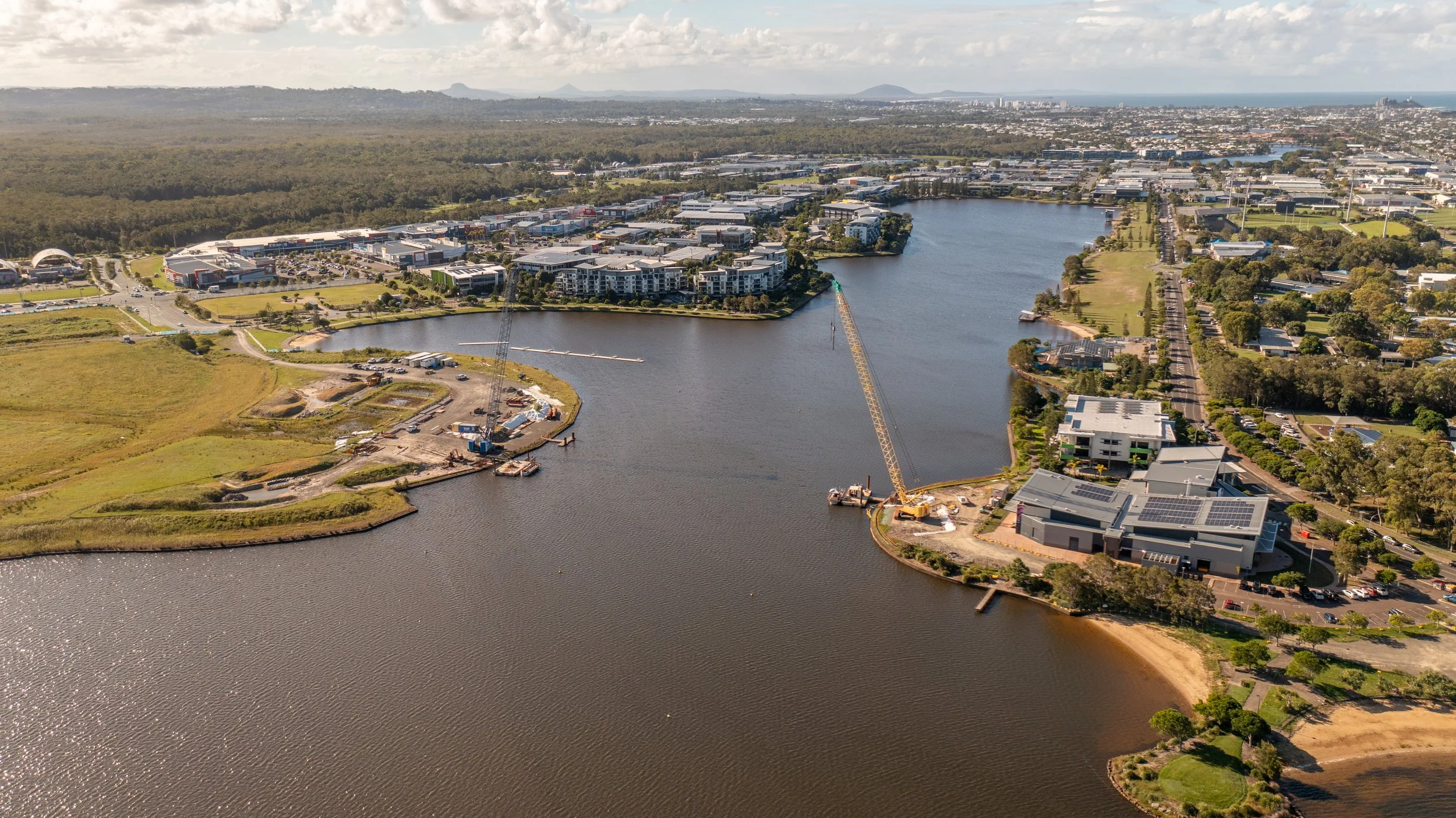 Aerial view of a waterway with construction equipment, buildings, and greenery along the shores, with a cityscape and mountains in the distance.