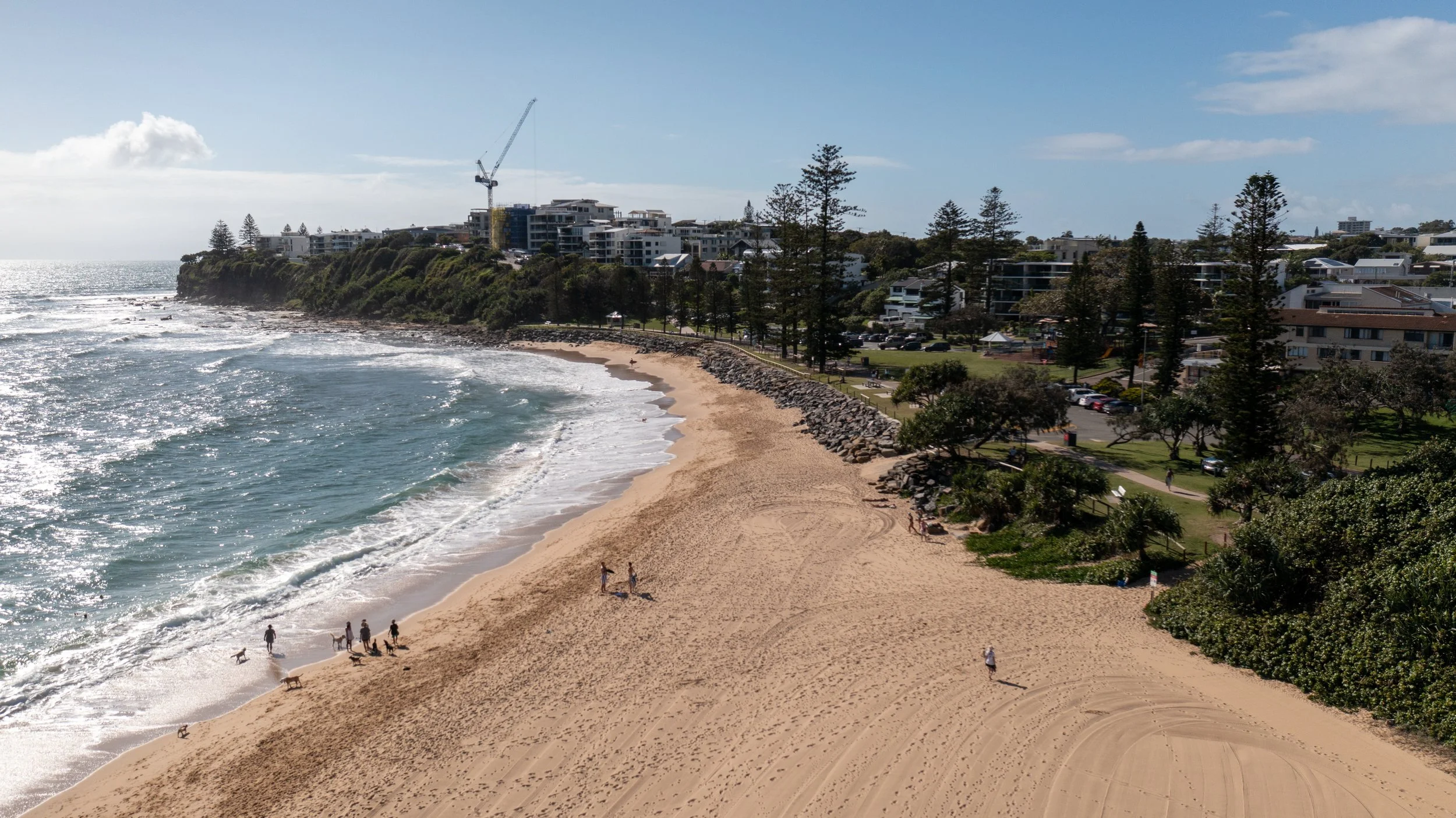 A scenic beach with golden sand, a few people and dogs walking, a rocky barrier, and ocean waves. In the background, there are modern buildings, trees, and a construction crane under a partly cloudy sky.