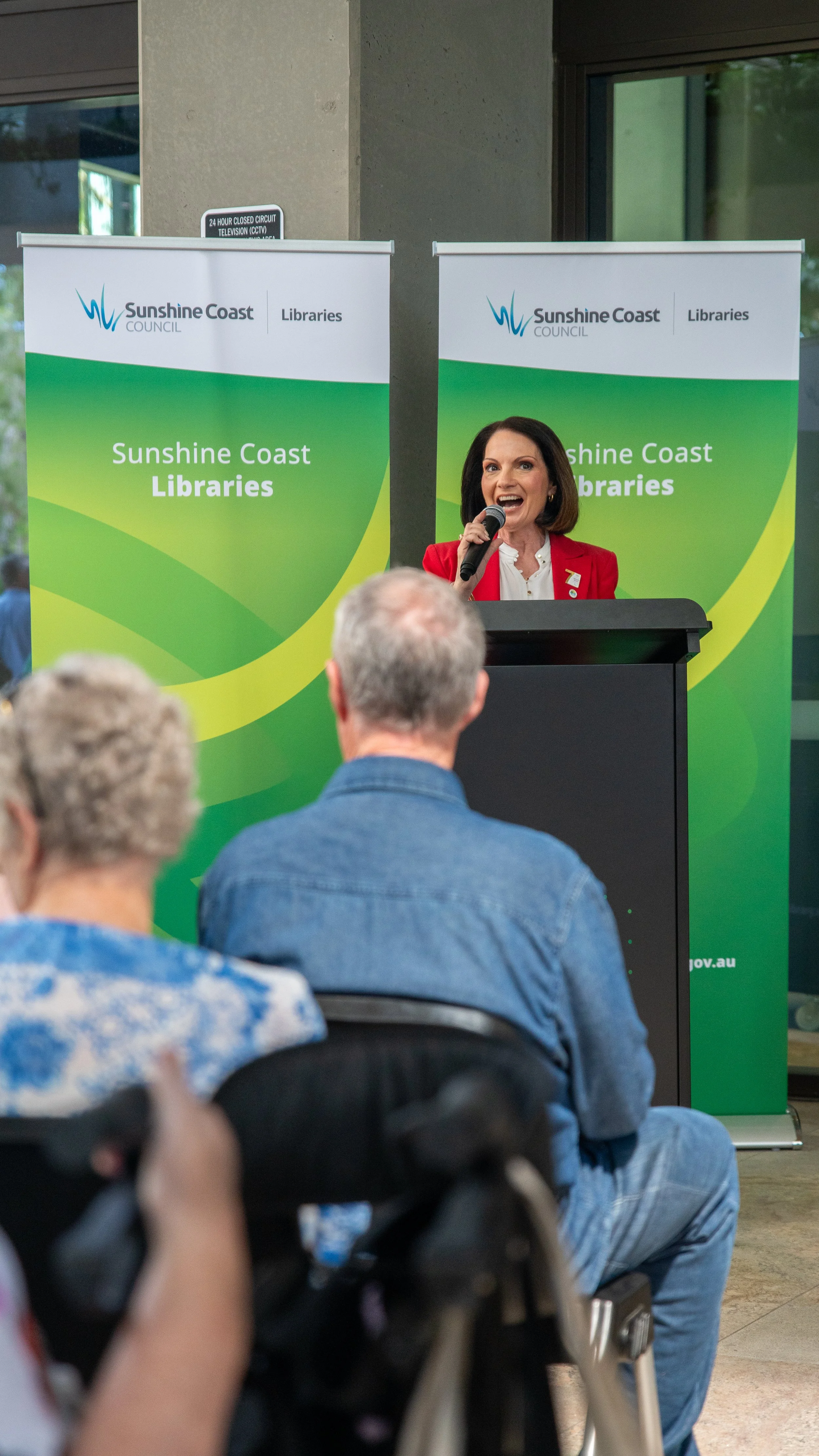 A woman standing at a podium, speaking into a microphone at a Sunshine Coast Libraries event. She is wearing a red blazer and has shoulder-length dark hair. In the background are two green banners with the Sunshine Coast Council logo and the words 'S