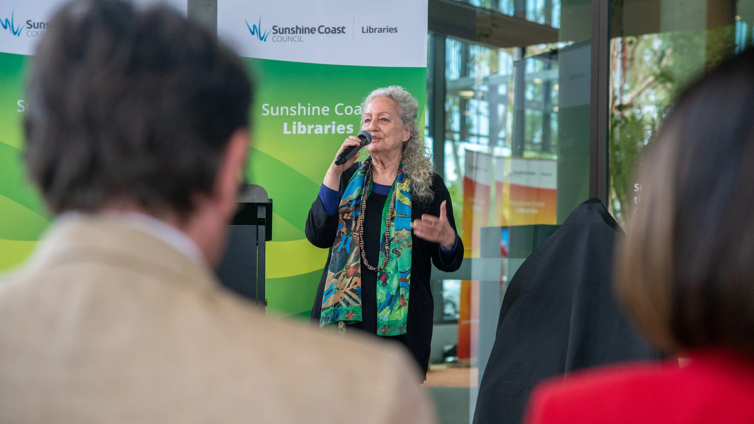 An elderly woman speaking into a microphone at a Sunshine Coast Libraries event, with a green and white backdrop and a glass building behind her, while an audience listens.