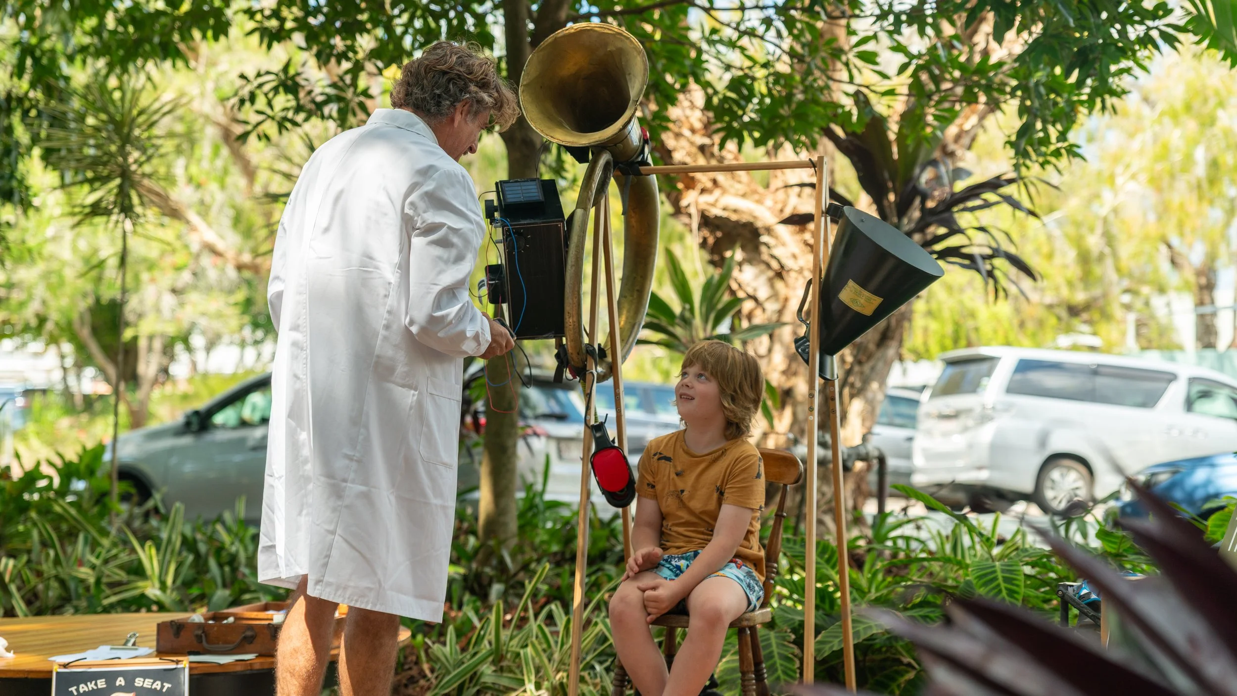 A man in a white lab coat is talking to a young boy sitting on a wooden chair outdoors, surrounded by green plants and trees. The man is holding a device connected to a large, brass horn and a black tube. The boy is looking up at the man, smiling.
