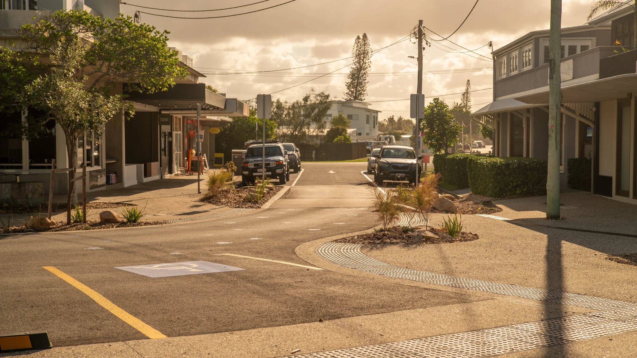 A small parking lot with several cars parked, a designated handicapped parking space, small landscaped islands with plants, trees, and rocks, surrounding a pedestrian walkway in a commercial area with buildings and power lines under a cloudy sky.