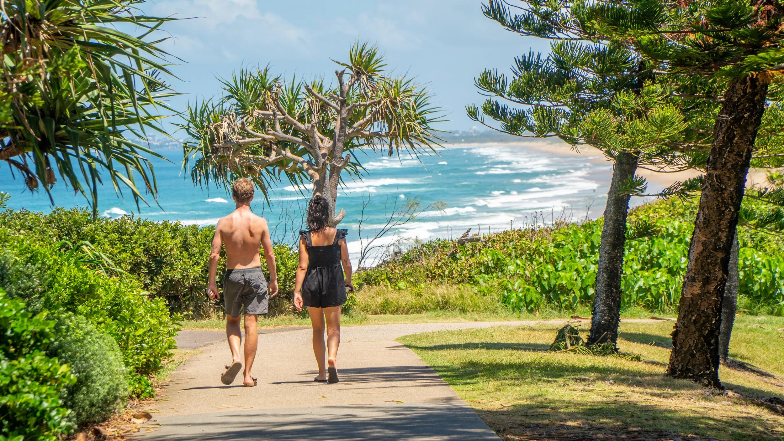 A couple walking along a sandy path near the beach, surrounded by lush green plants and trees with the ocean in the background.