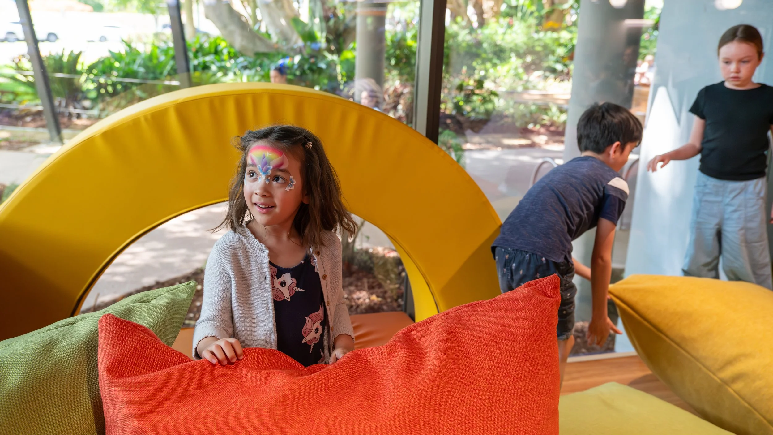 A young girl with face paint of a rainbow, unicorn, and flowers, standing behind orange and green cushions in a room with large glass windows and plants outside. Two boys are nearby, one bent down and the other standing, in a play area.