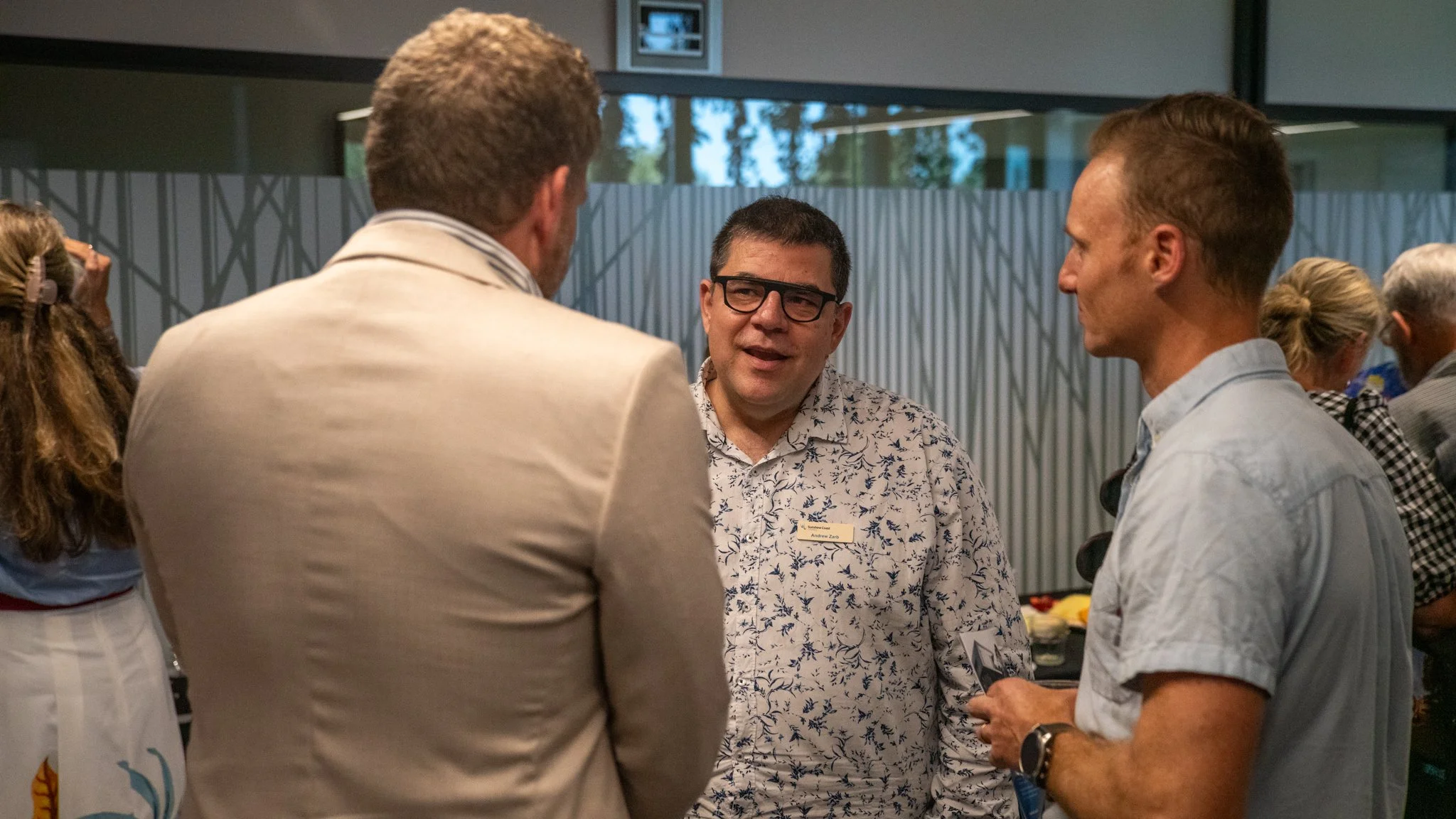 Three men are engaged in a conversation at a social event, with a woman in the background. The man in the middle has glasses and is wearing a patterned shirt, while the other two men have lighter shirts. They are in a room with large windows and mode