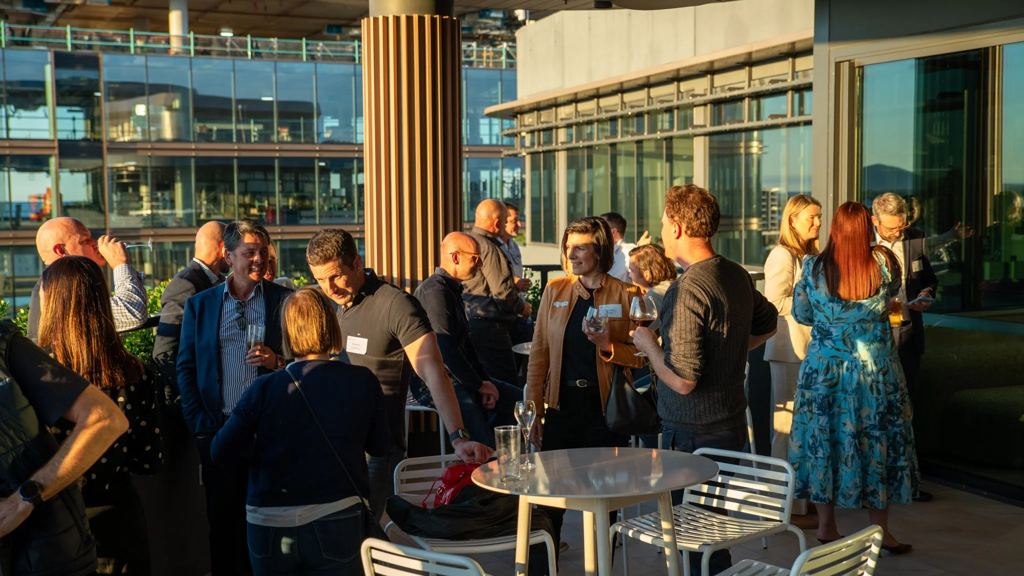 People socializing at an outdoor rooftop event during sunset, with glass buildings in the background.