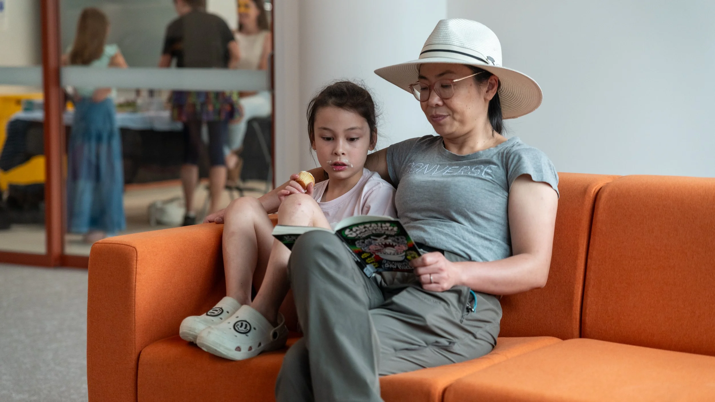 A woman and a young girl sitting on an orange sofa in a waiting area, reading a comic book together. The woman is wearing a wide-brimmed hat and glasses, and the girl has a snack in her mouth.