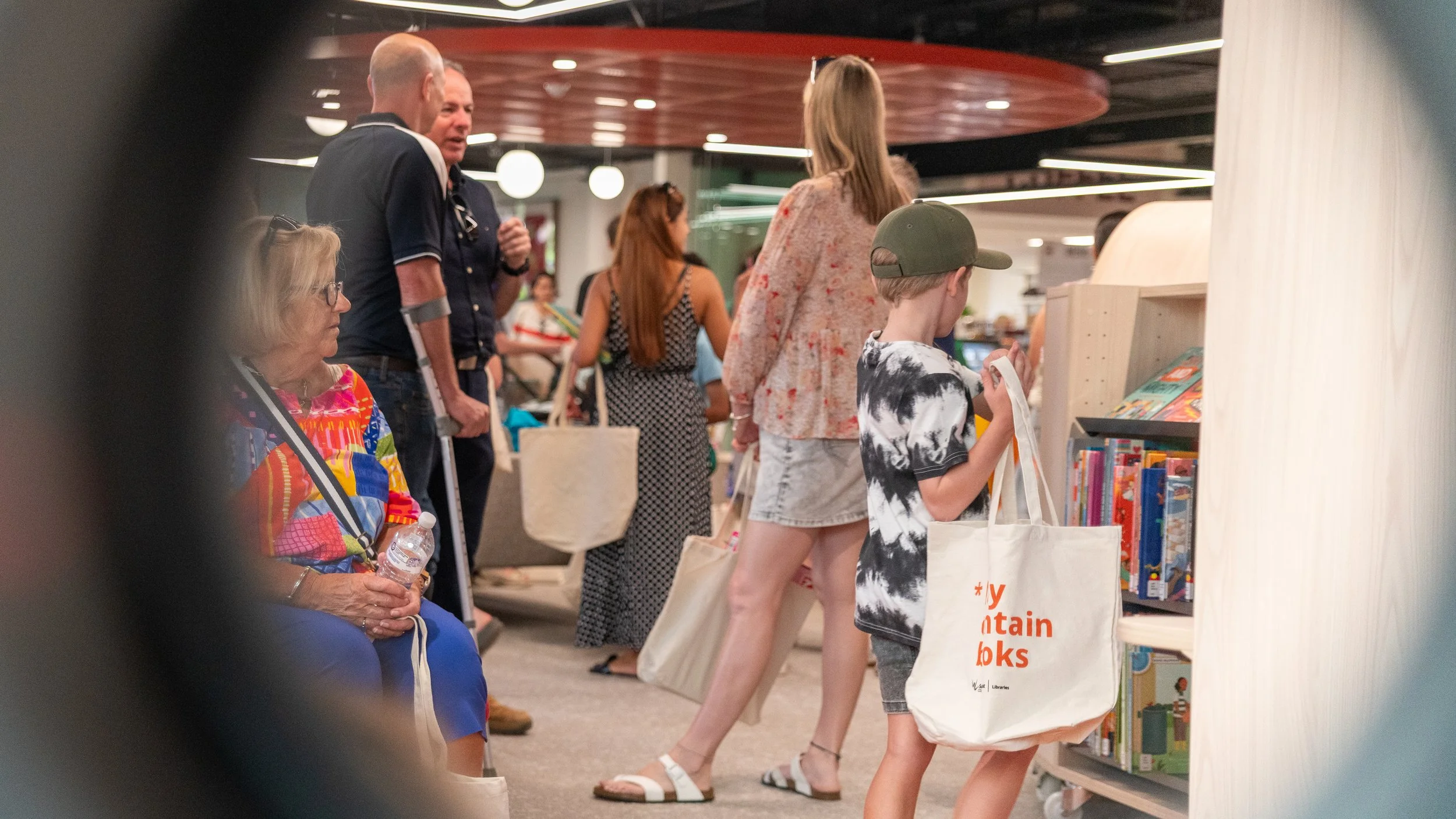 People shopping at a bookstore, with a woman sitting with a water bottle, multiple customers browsing books, and a young boy looking at books on a shelf.