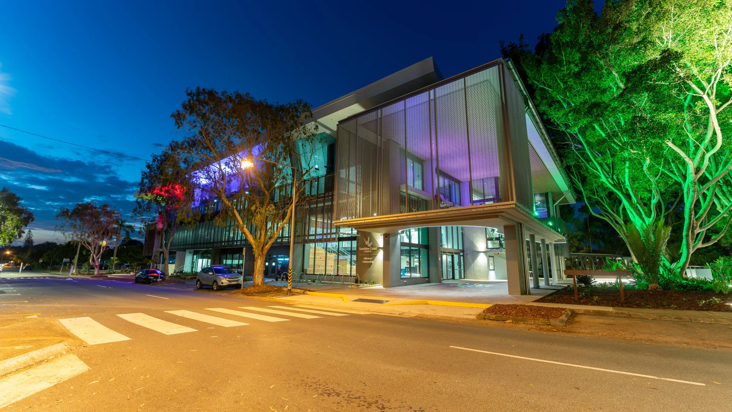 Modern multi-story building with glass exterior illuminated with colorful lighting, surrounded by trees, and located on a city street at dusk.