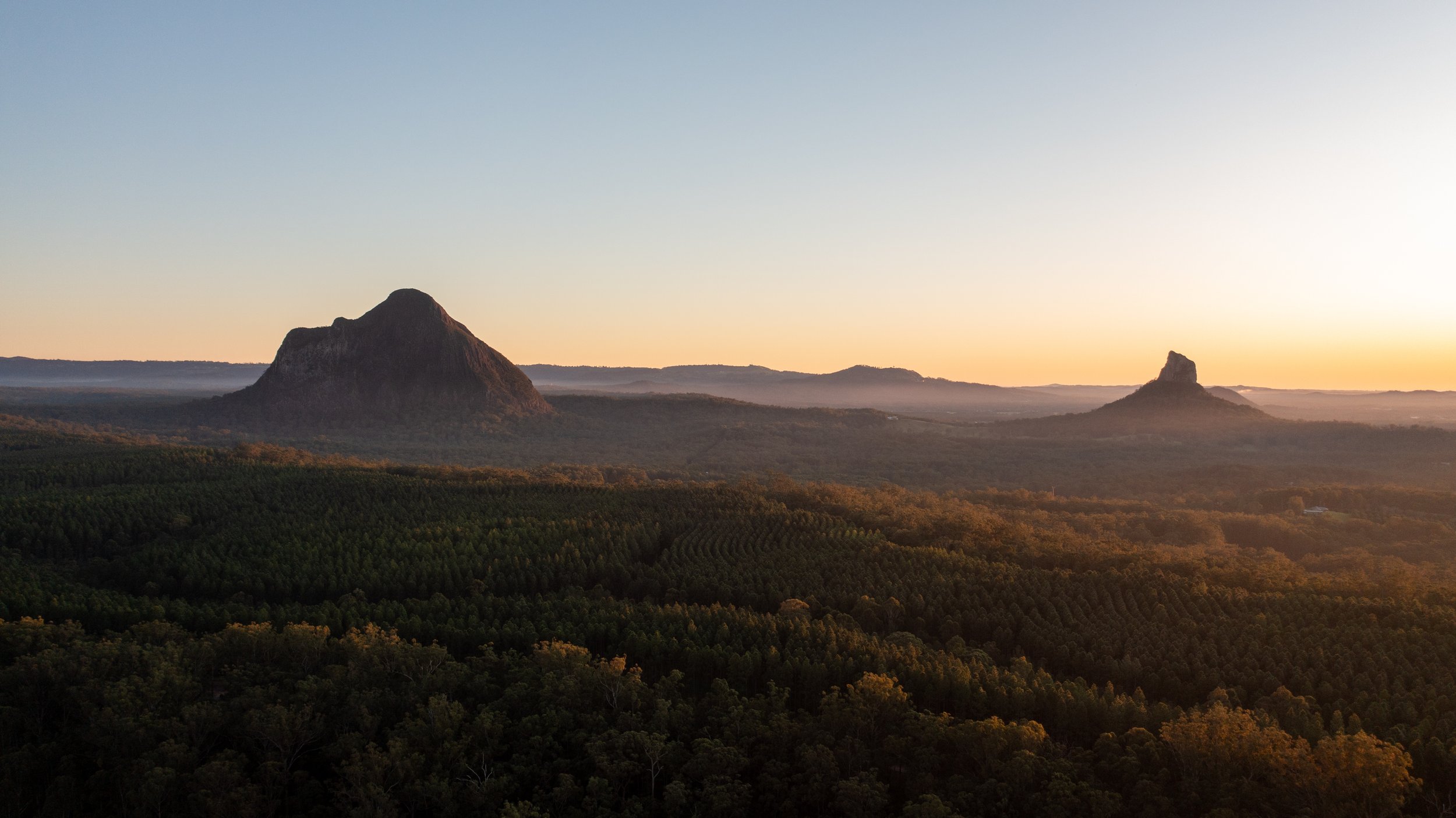Sunrise over forested hills with two prominent mountain formations in the distance.