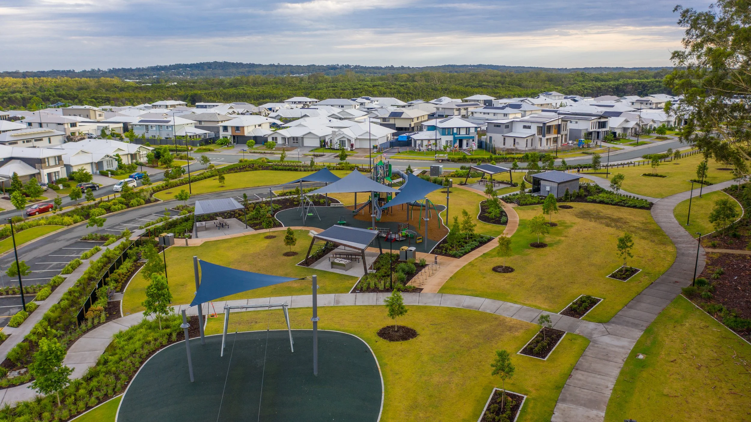 Aerial view of a residential neighborhood with neatly arranged houses with white roofs, a park area with walking paths, shade sails, a playground, trees, benches, parking spaces, and a surrounding green landscape under a cloudy sky.