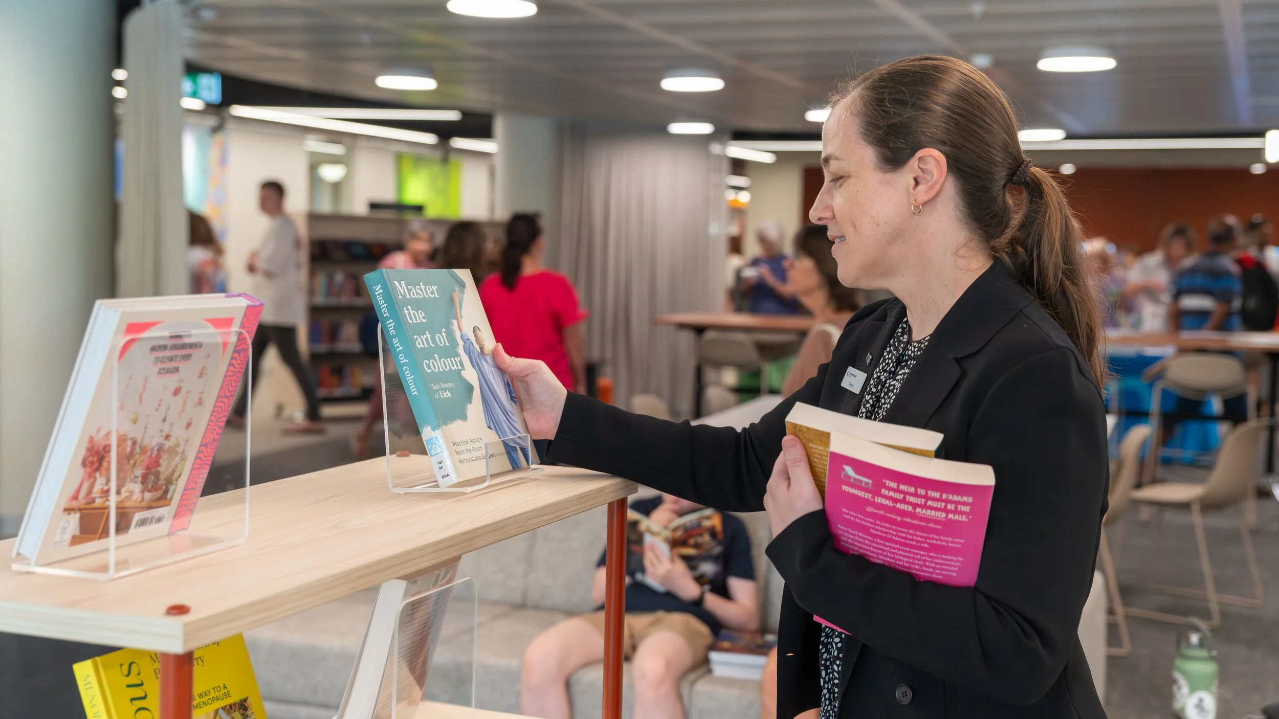 A woman with brown hair tied back, wearing a black blazer, browsing books at a public place like a library or bookstore, with several people in the background.