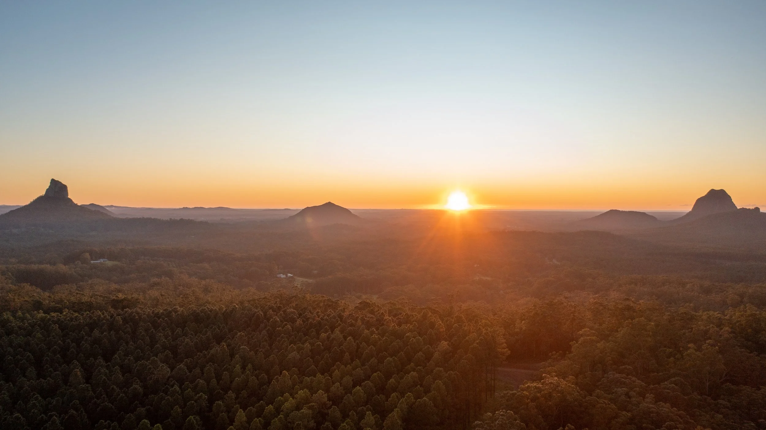 Sunset over a forested landscape with three prominent rocky formations in the distance.