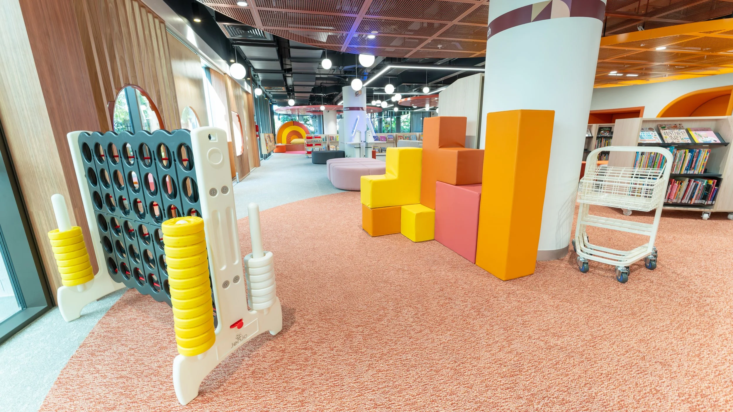 Children's indoor play area with Connect Four game, colorful foam steps, bookshelf, and a lounging area with pink seating.