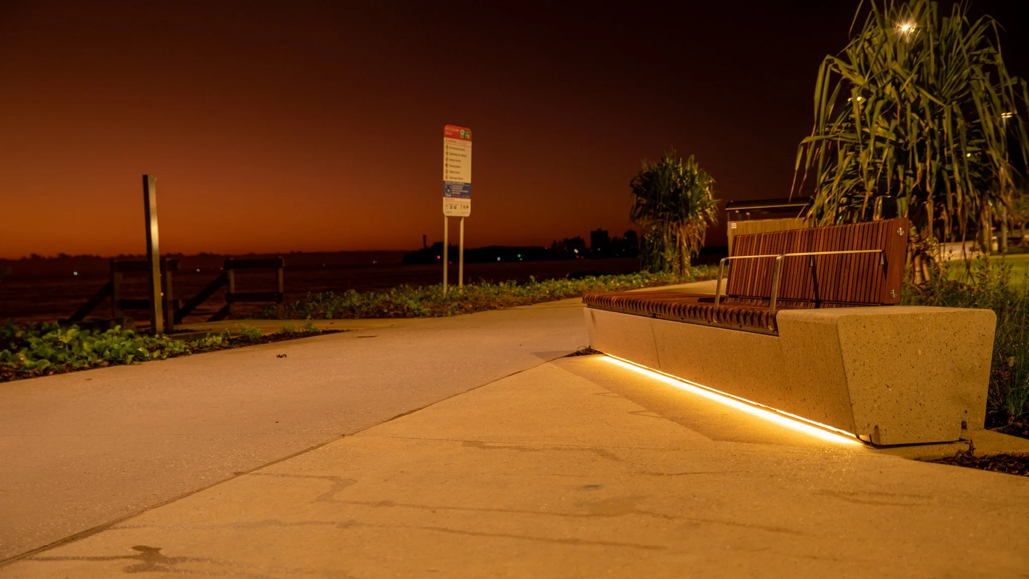 An empty park bench illuminated by ground lighting at night, with a sunset sky and waterfront in the background.