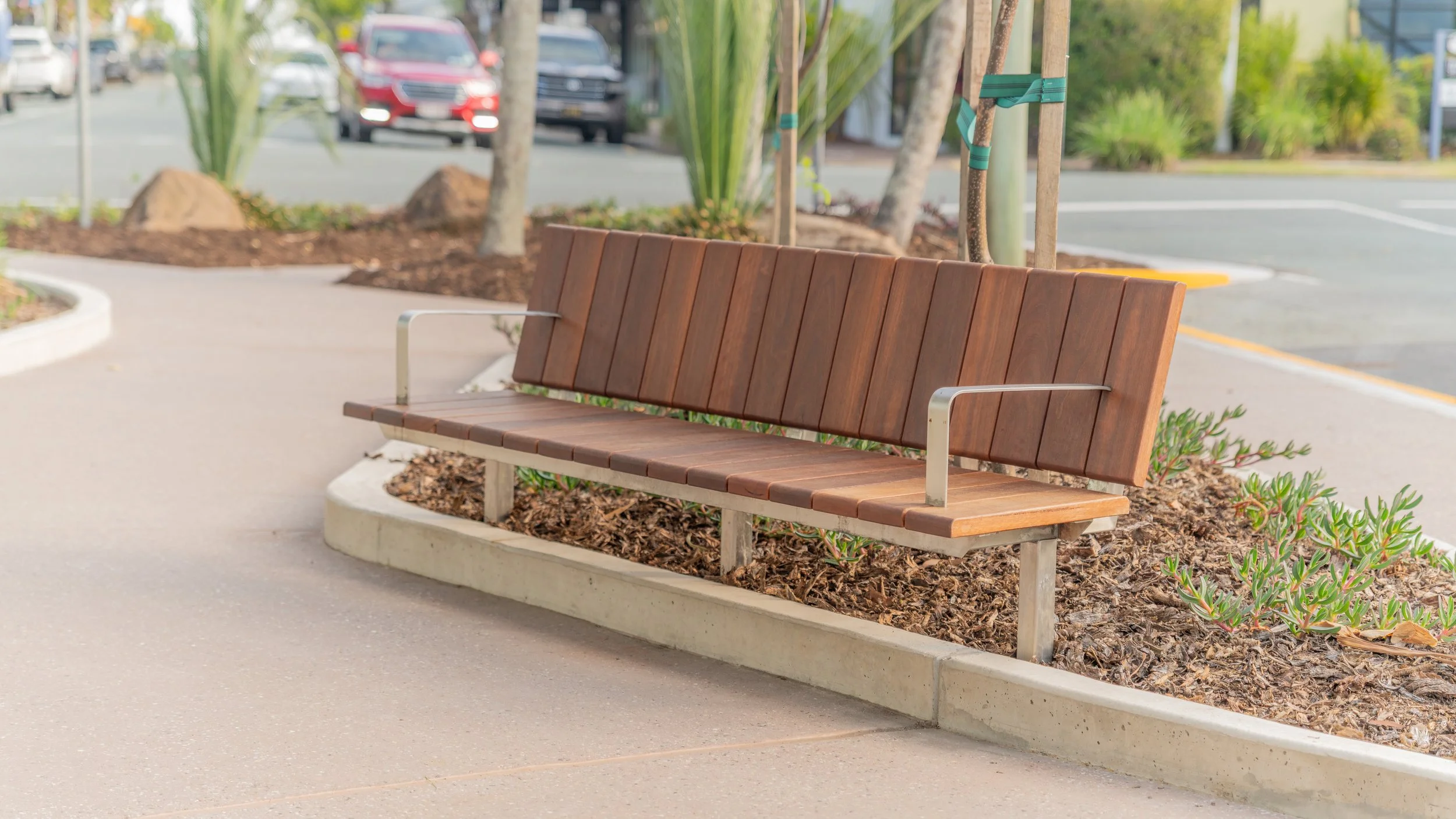 Wooden park bench with metal armrests on a concrete sidewalk next to landscaped area with mulch and plants, in a parking lot with cars and trees in the background.