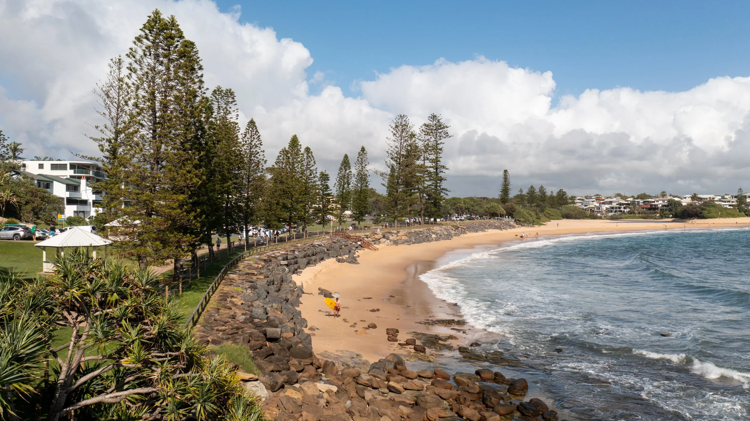 Beach with sandy shore, rocks along the edge, and waves crashing onto the sand. Tall trees line the walkway parallel to the beach, and there are buildings in the background. People are walking and playing on the beach on a partly cloudy day.