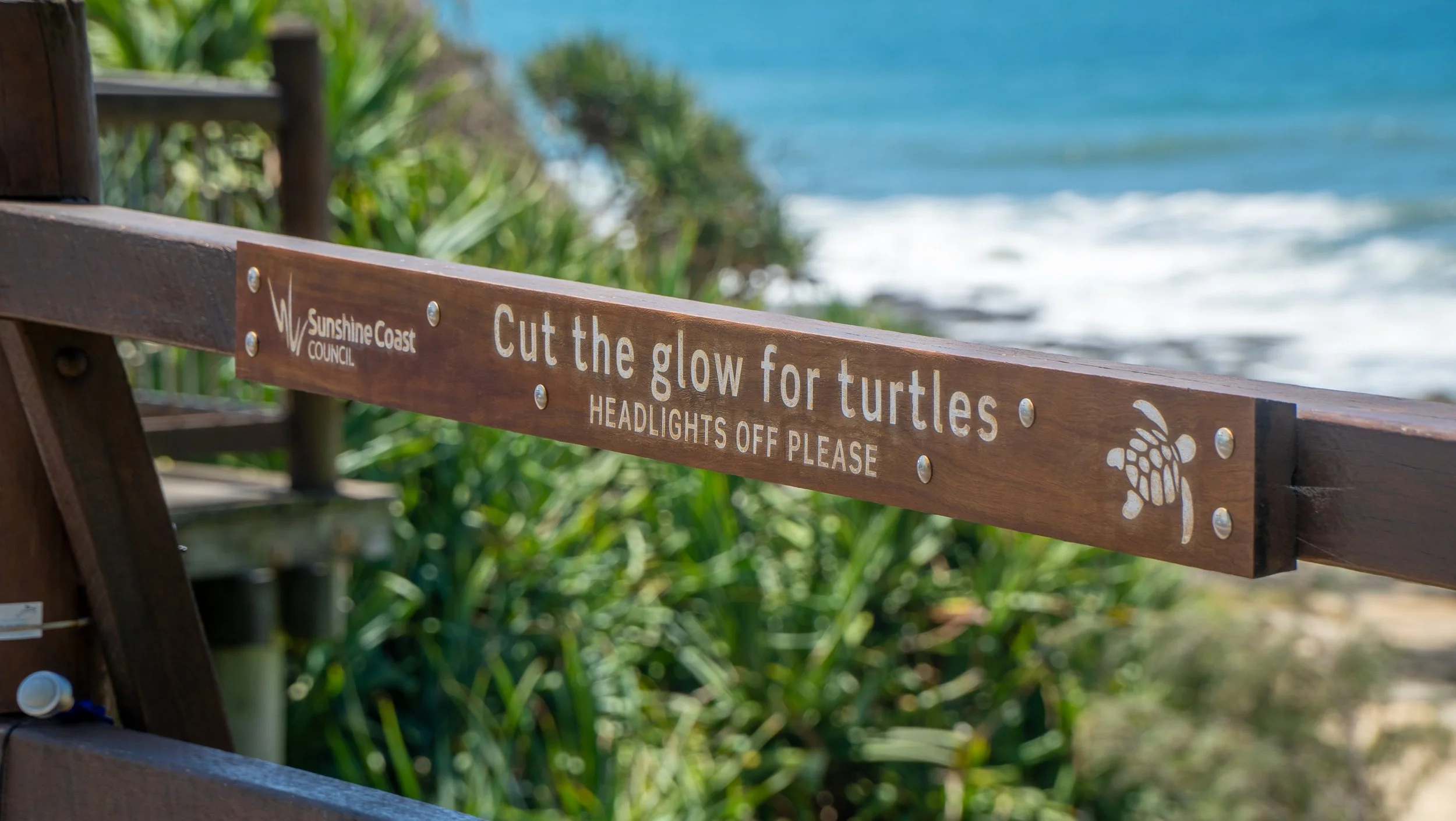 A wooden sign on a railing at the beach with the message: 'Cut the glow for turtles. HEADLIGHTS OFF PLEASE,' and a turtle illustration, with ocean and greenery in the background.