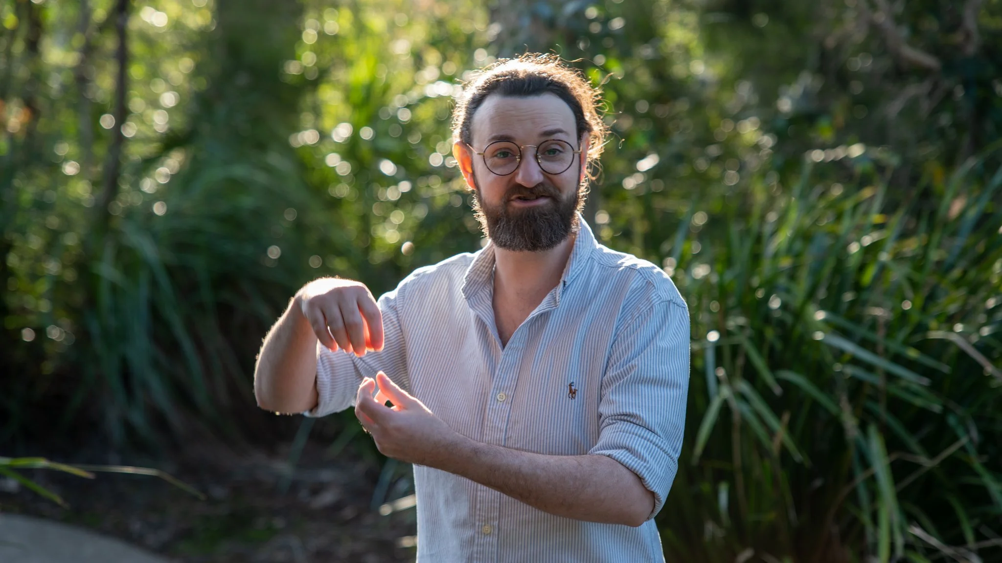 Man with glasses and a beard standing outdoors in a forested area with green foliage, wearing a light-colored, striped button-up shirt, making a gesture with his hands that resembles adjusting or showing something.